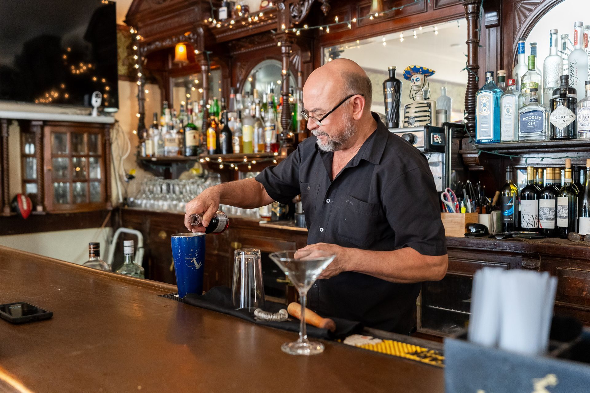 Bartender making a martini in a dimly lit bar; he's bald and smiling; bottles and glasses are on the shelves behind him.