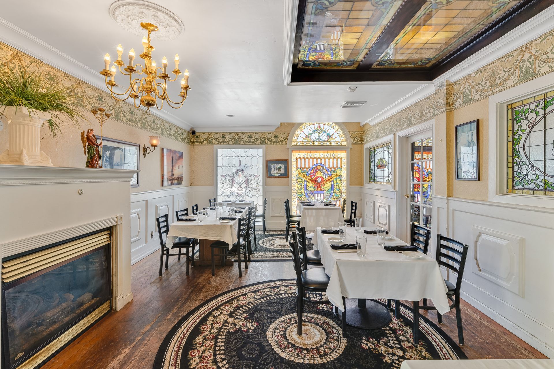 Restaurant interior: tables with white linens, black chairs, fireplace, stained glass windows, chandelier, patterned carpet.