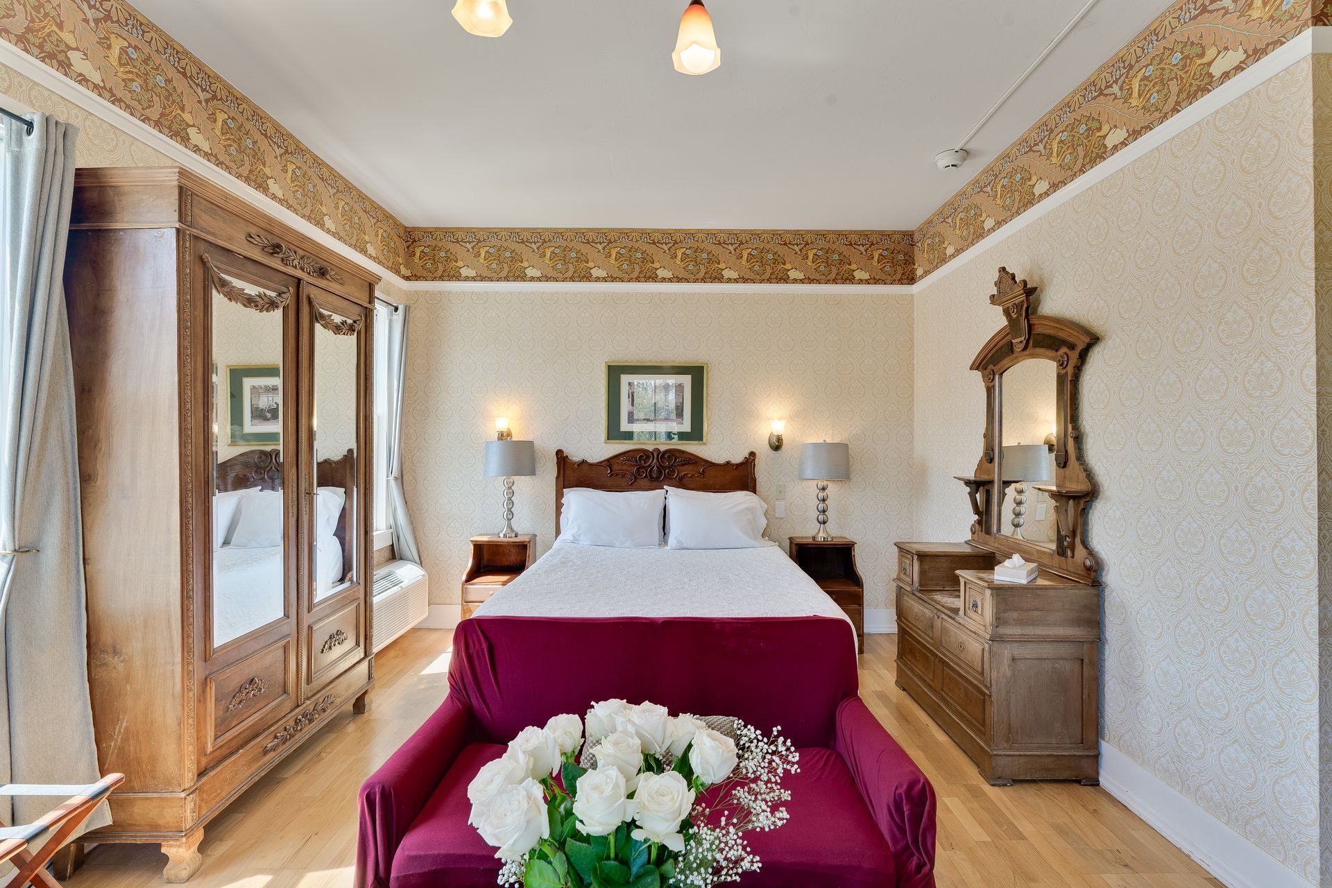 Bedroom with antique furniture, including a large armoire, dresser with mirror, and a maroon chair with white roses.
