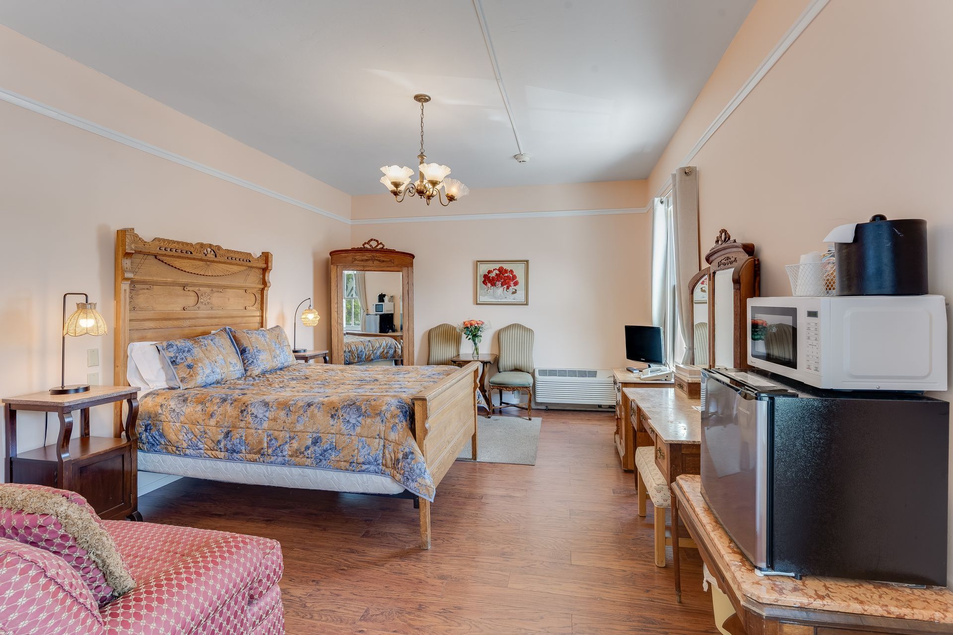 Bedroom with bed, microwave, mini-fridge, antique furniture. Pink walls, floral bedspread, and a sofa.