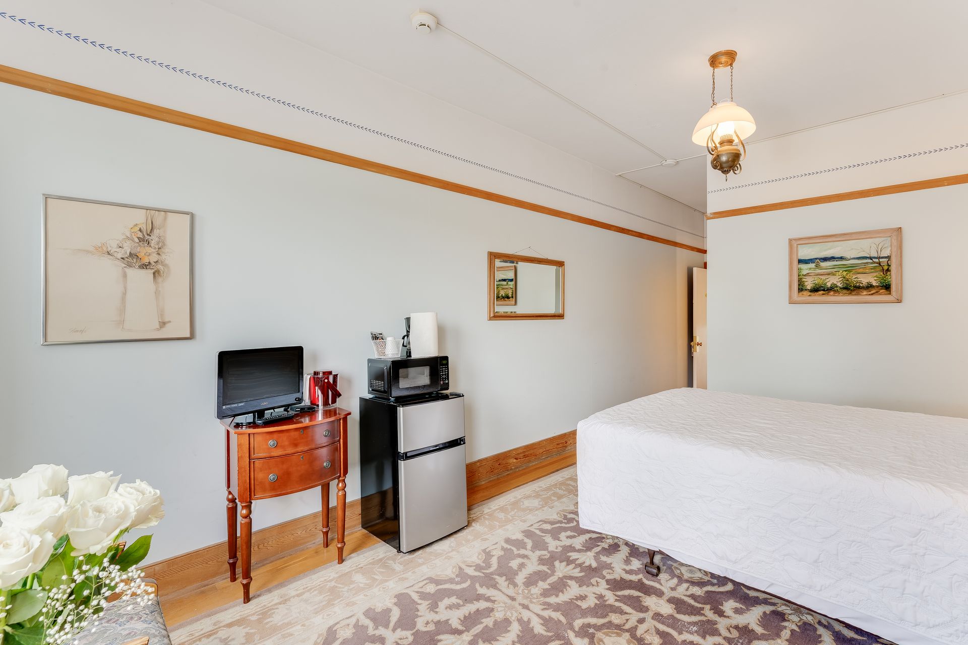 Bedroom with bed, TV, microwave, fridge, artwork, and flowers.