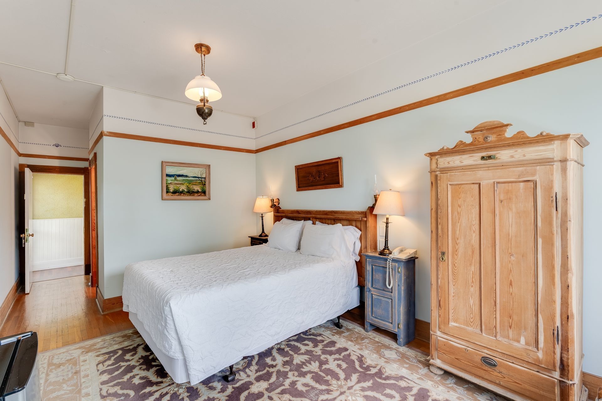 Bedroom with a bed, wooden wardrobe, and bedside tables, light blue walls and a patterned rug.
