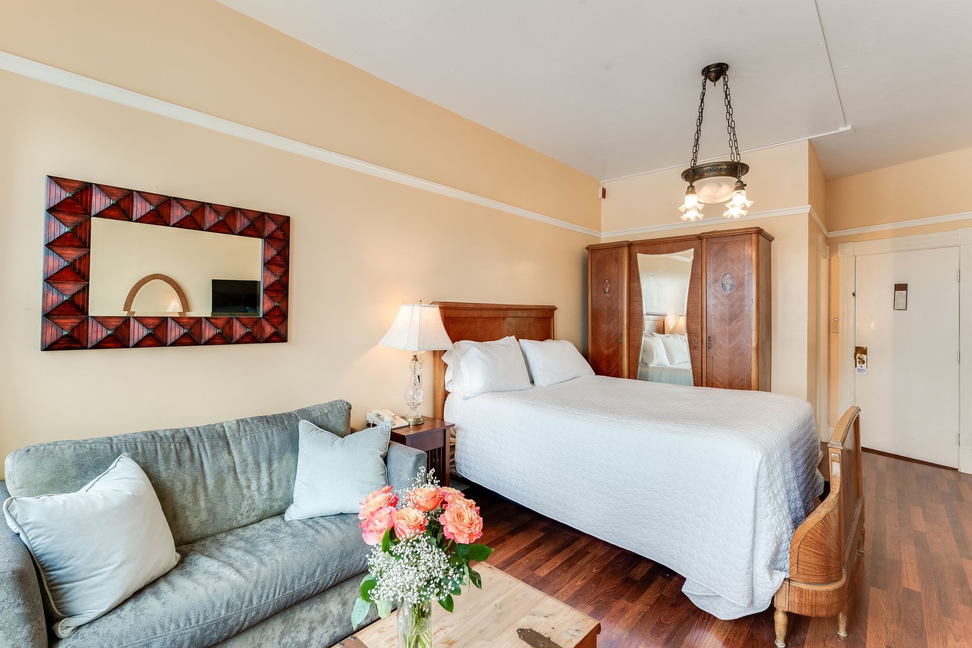 Cozy hotel room with a bed, sofa, mirror, and wardrobe. Warm colors and wooden floors.
