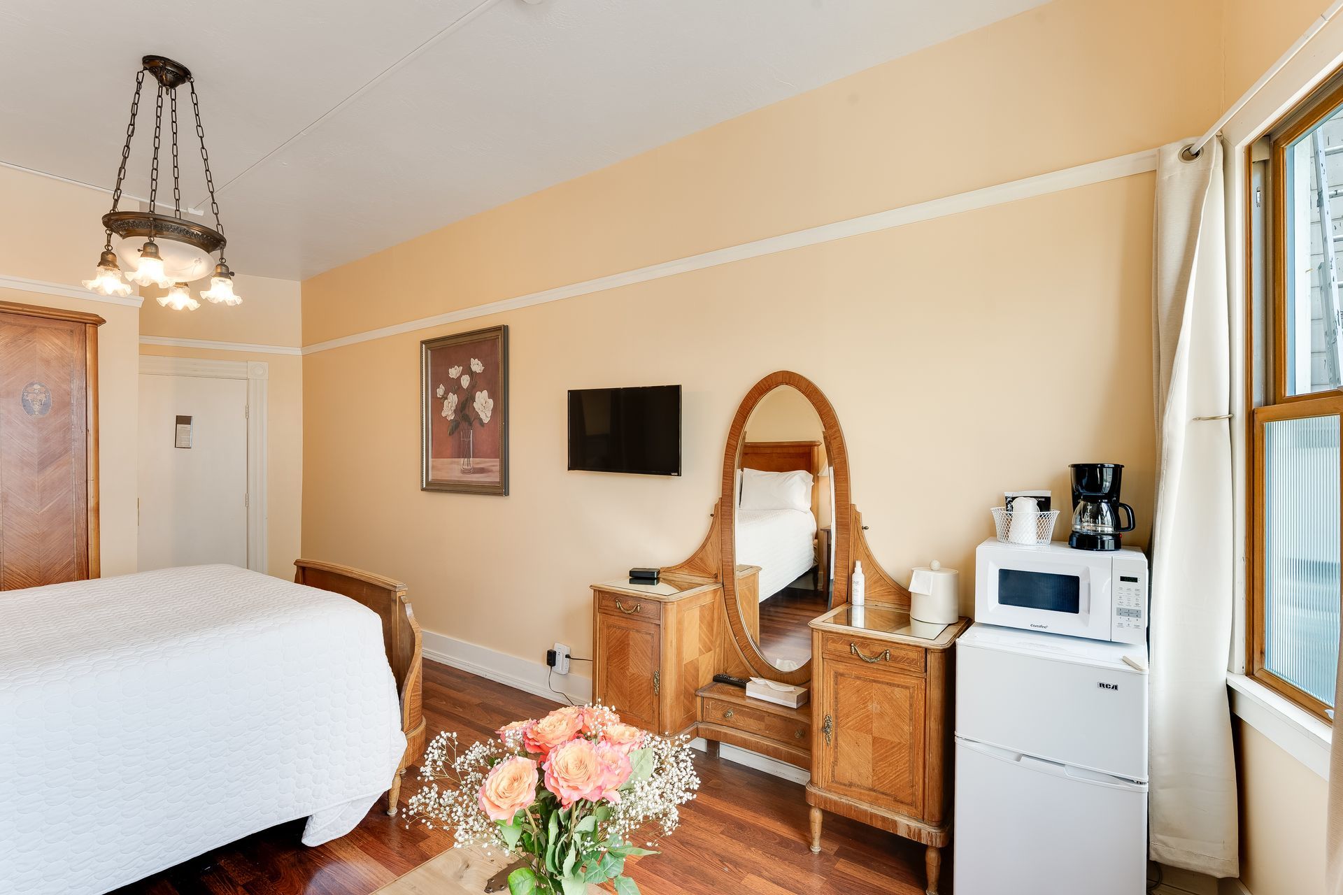 Cozy hotel room: bed, antique vanity, TV, microwave, mini-fridge, roses, and hanging light.