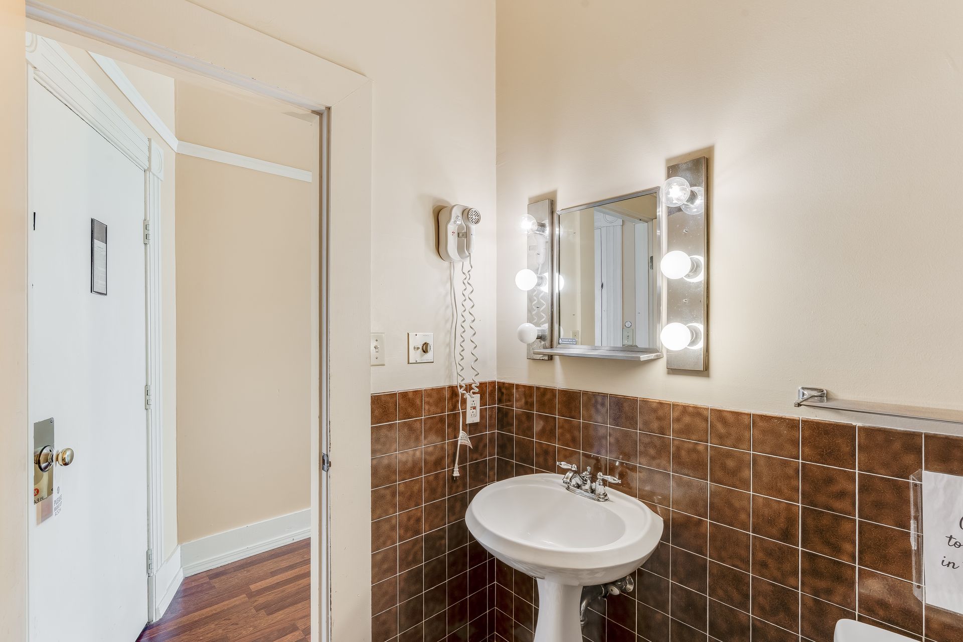 Bathroom with white sink, brown tile, and a mirror with lights. Doorway on the left.