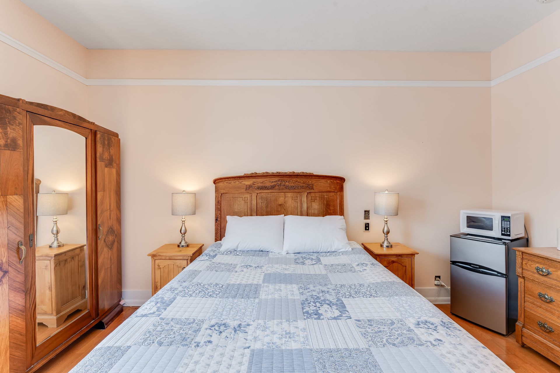 Bedroom with wood furniture, bed, nightstands, armoire, microwave, mini-fridge. Light pink walls.