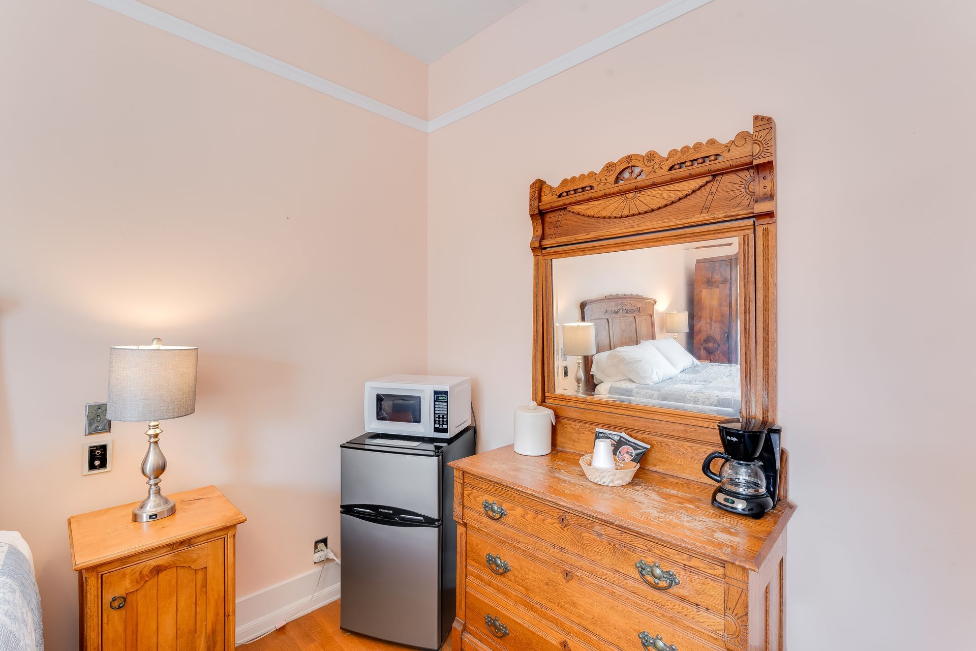 A bedroom corner with a dresser, mirror, mini-fridge, microwave, lamp, and coffeemaker.