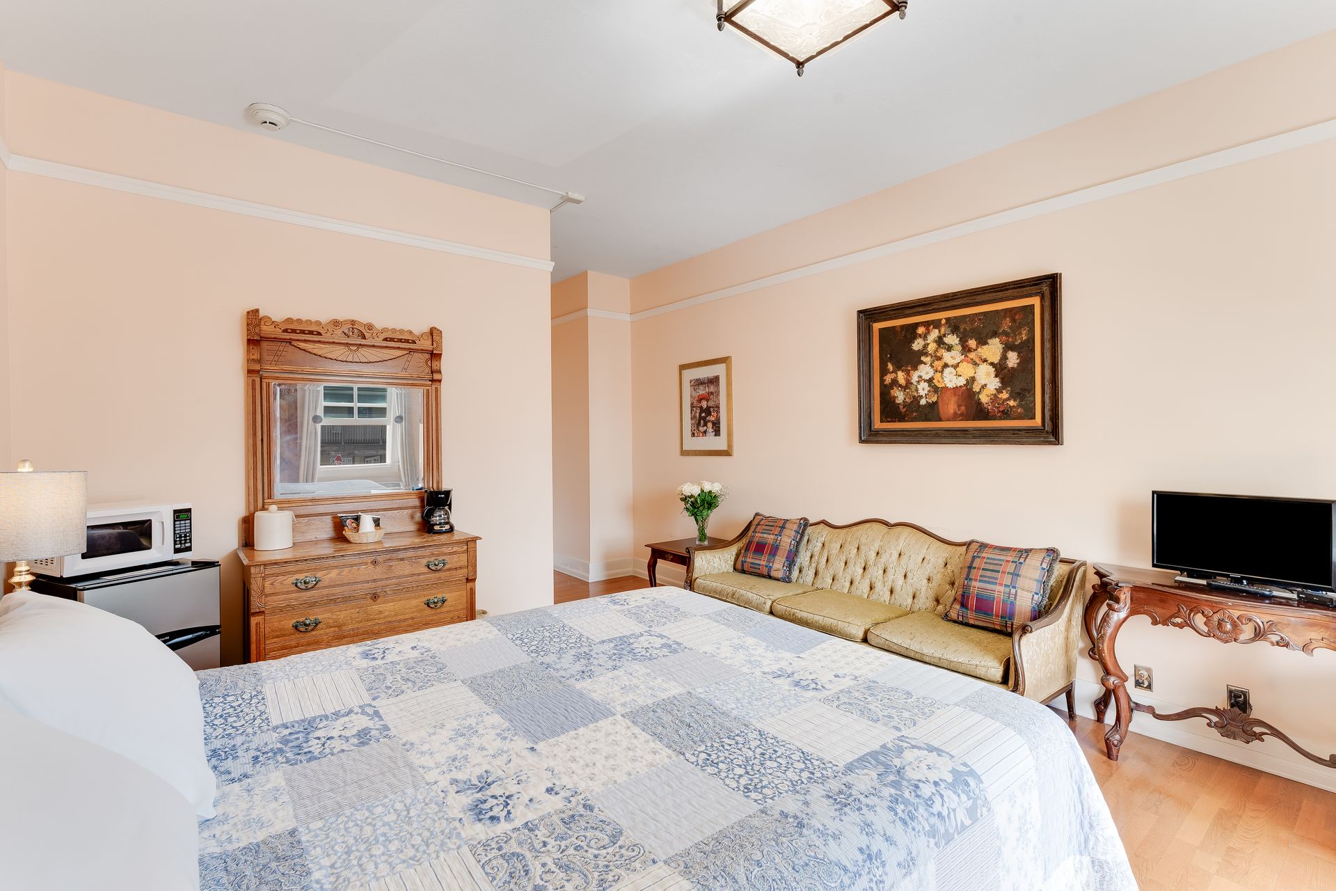 Bedroom with bed, couch, dresser, TV, and artwork. Soft peach walls and wood floors.
