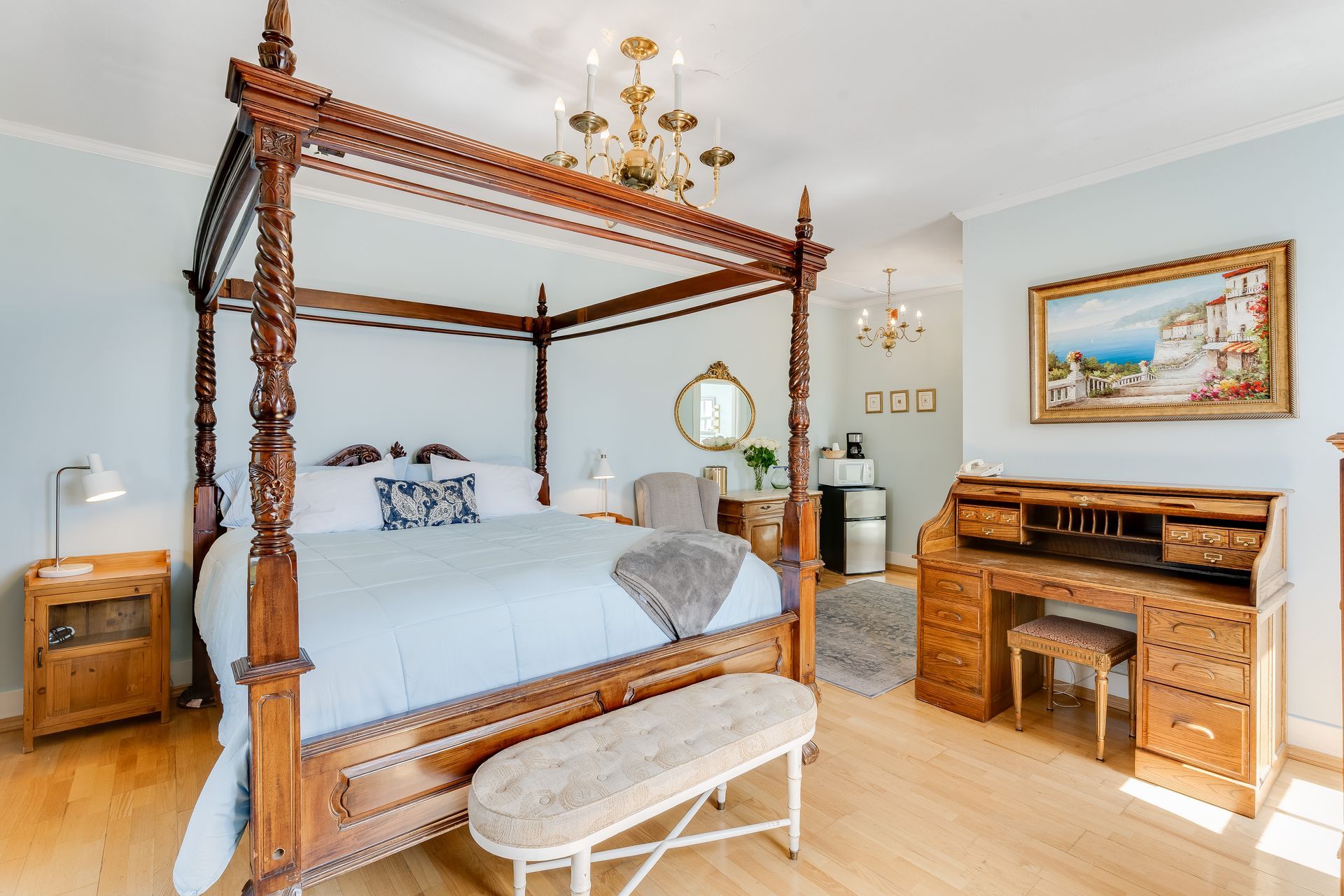 Bedroom with a four-poster bed, antique desk, small fridge, and a painting. Soft blue walls and light wooden floors.