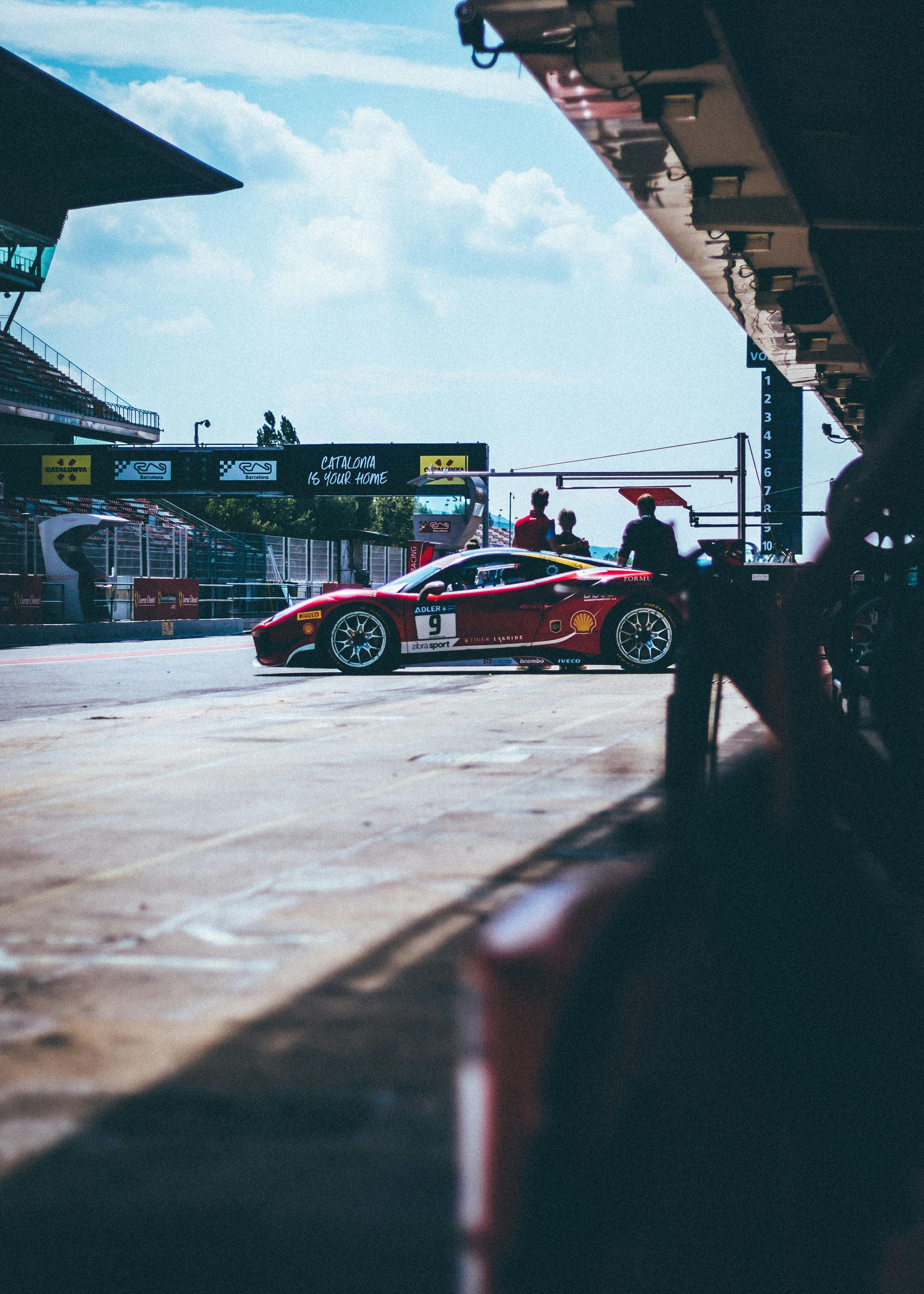 Race car in pit lane, red with yellow accents, parked near crew members and garages, blue sky overhead.