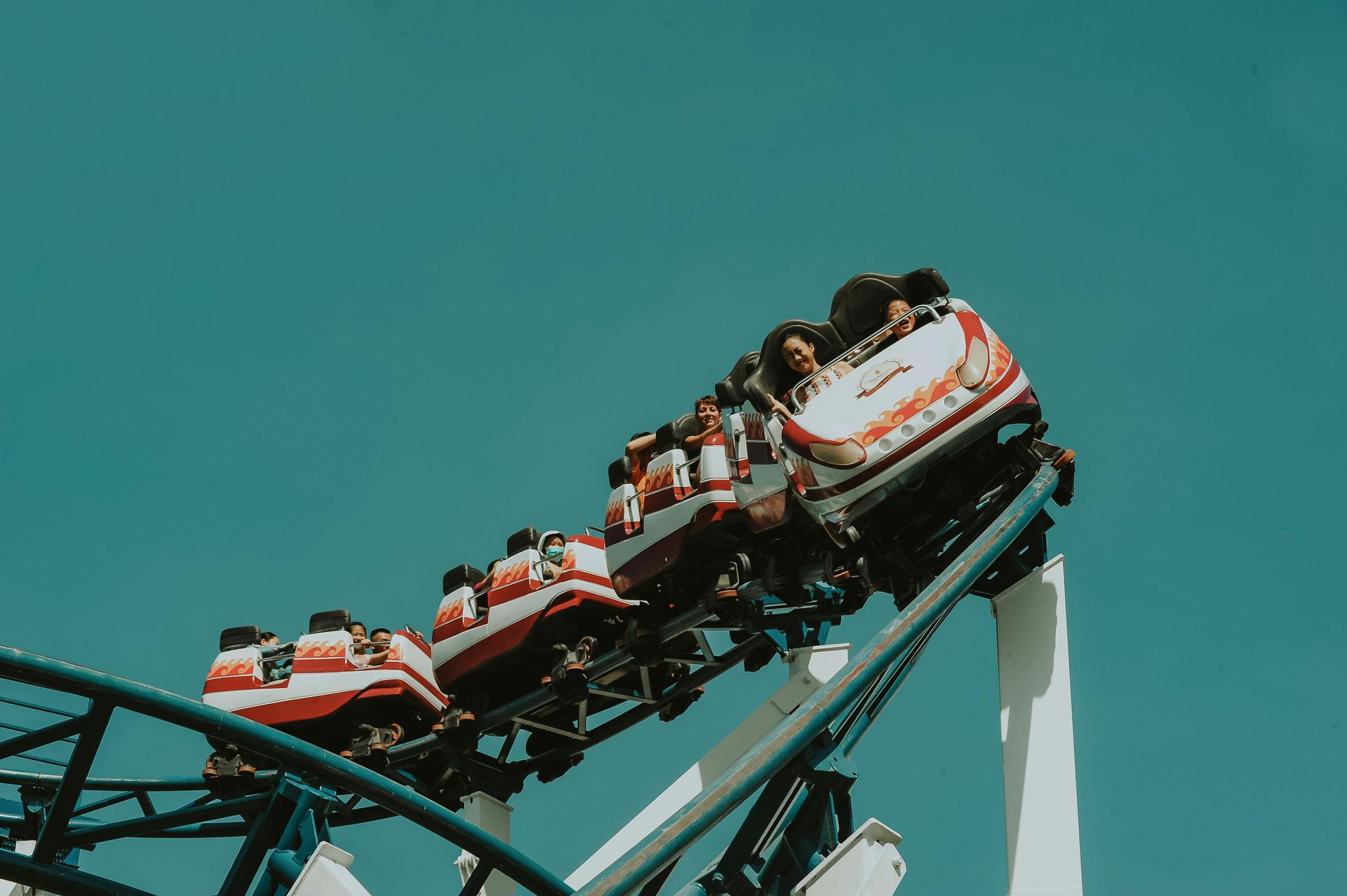 Roller coaster car ascending a blue track, people with excited expressions against a teal sky.