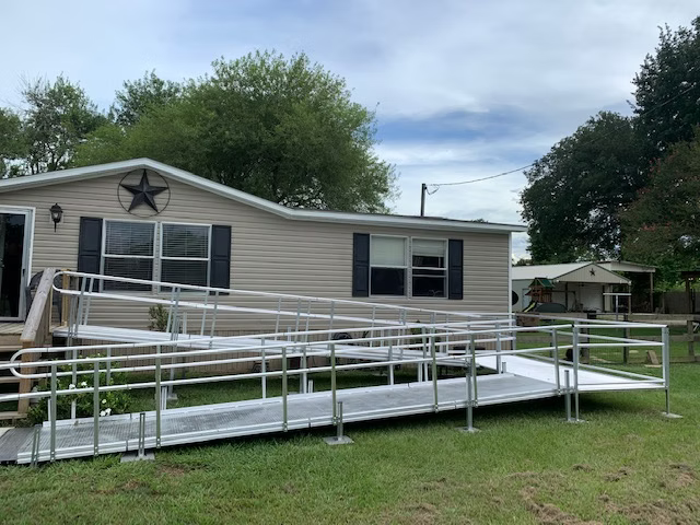 A tan mobile home with a silver wheelchair ramp extending from the entrance, set on a green lawn.