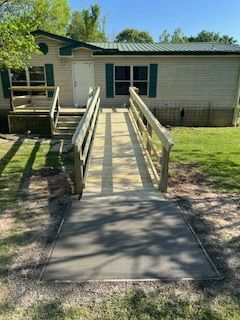 Exterior of a beige mobile home with a wooden ramp leading to the front door. Green shutters and a metal roof.