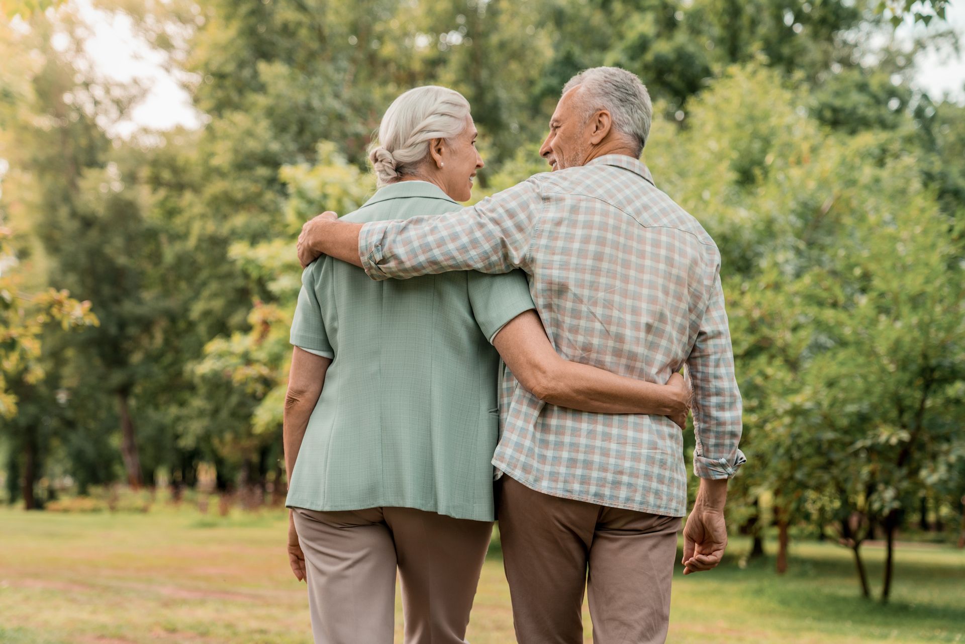 Two people walk away from the camera in a park with their arms around each other, wearing casual shirts and trousers.