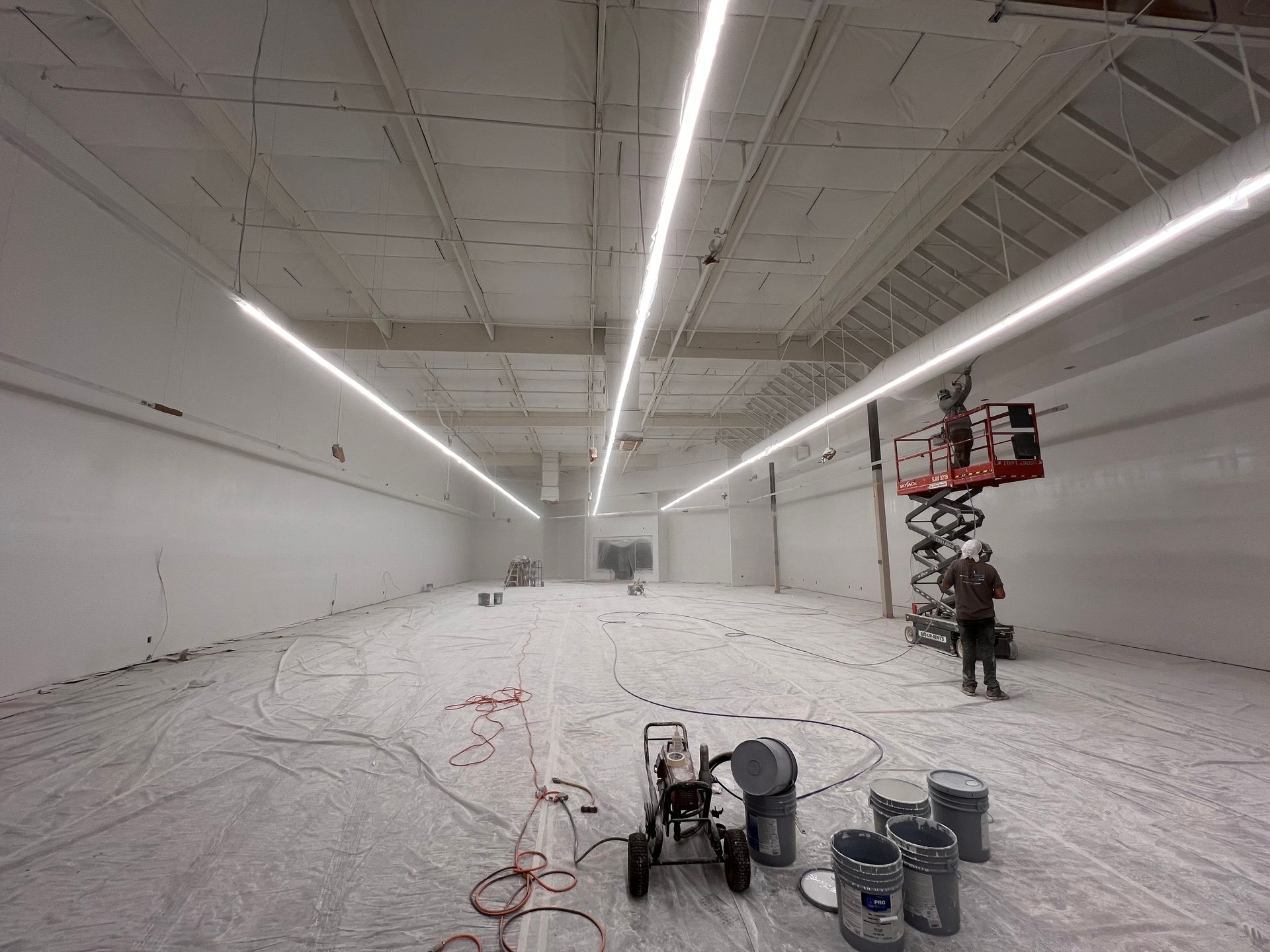 Man sanding a drywall ceiling with a long-handled sander. He wears a white shirt and gloves in a room with green drywall.