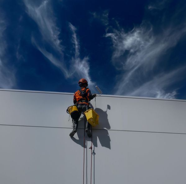 Rope access technician on a white building, cleaning, bright blue sky with clouds.