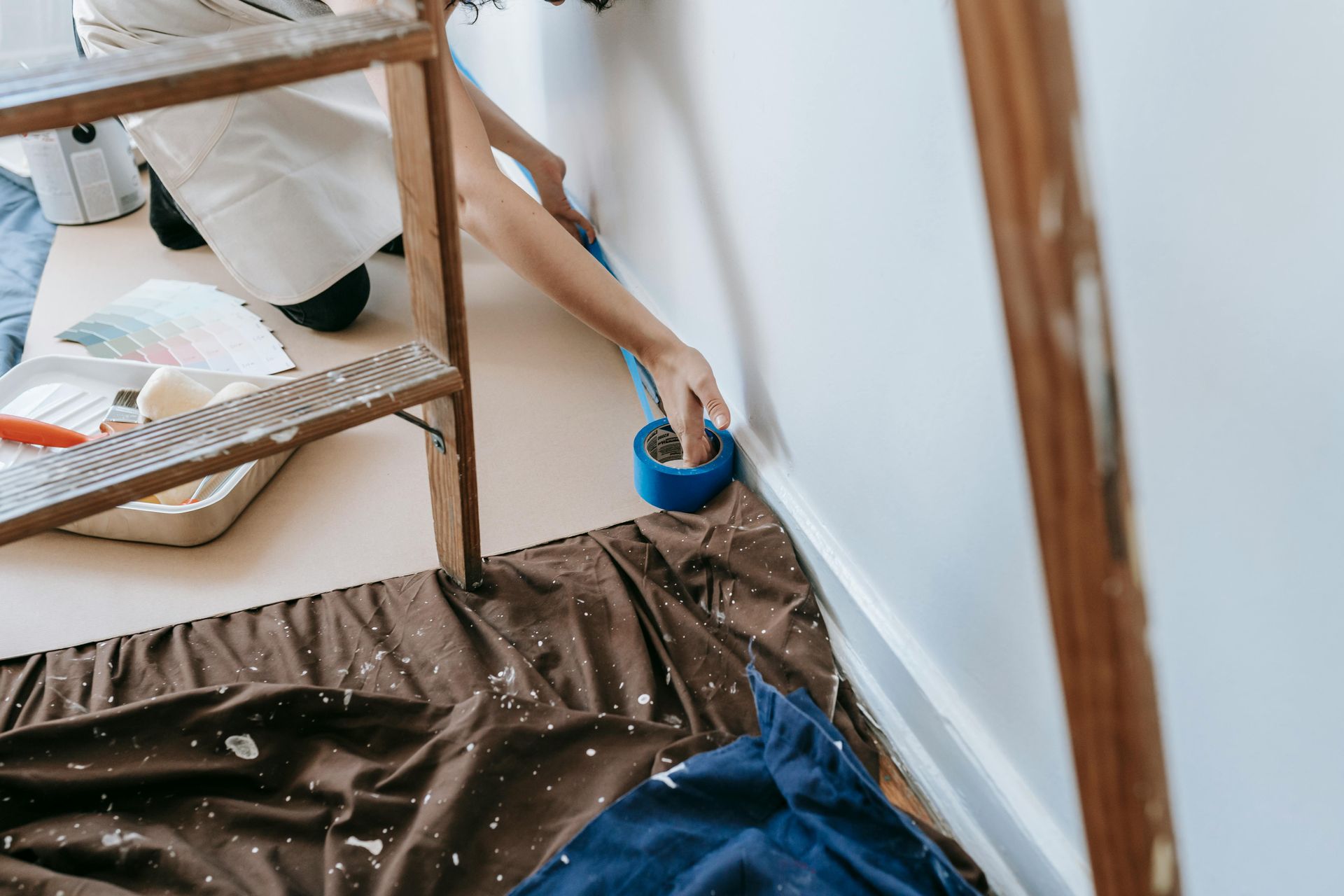Person taping a white wall while painting, with a ladder, drop cloth, and paint supplies.