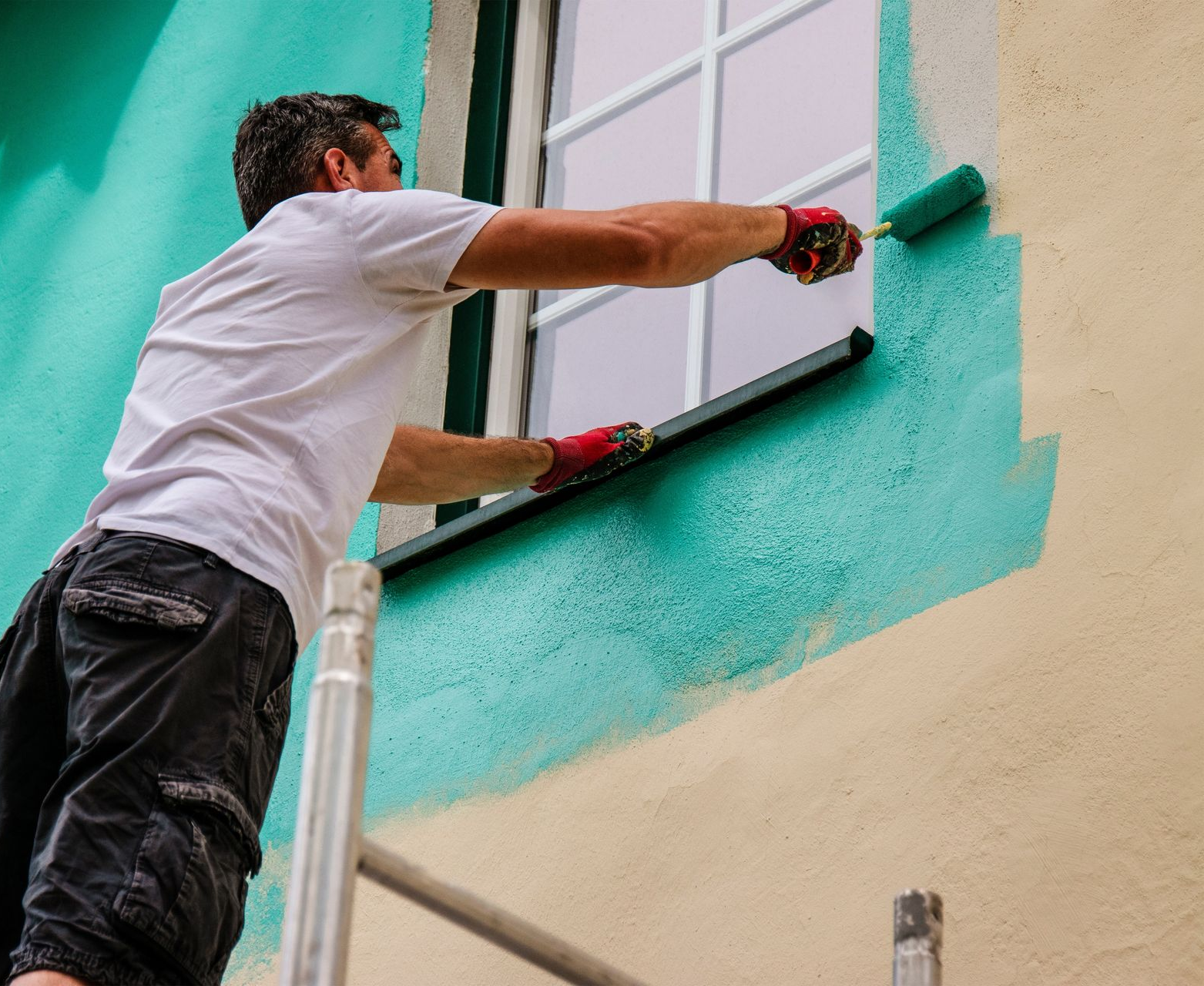 Man painting a turquoise wall around a window, using a roller, with a light tan wall.