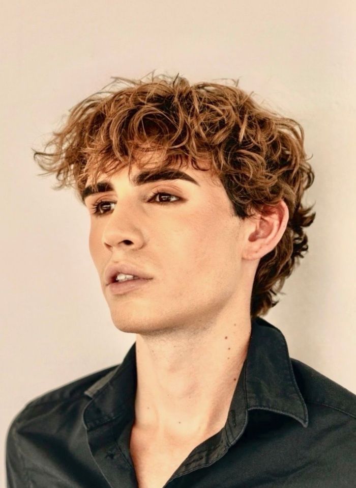 Portrait of a person with curly hair wearing a dark shirt against a plain beige background