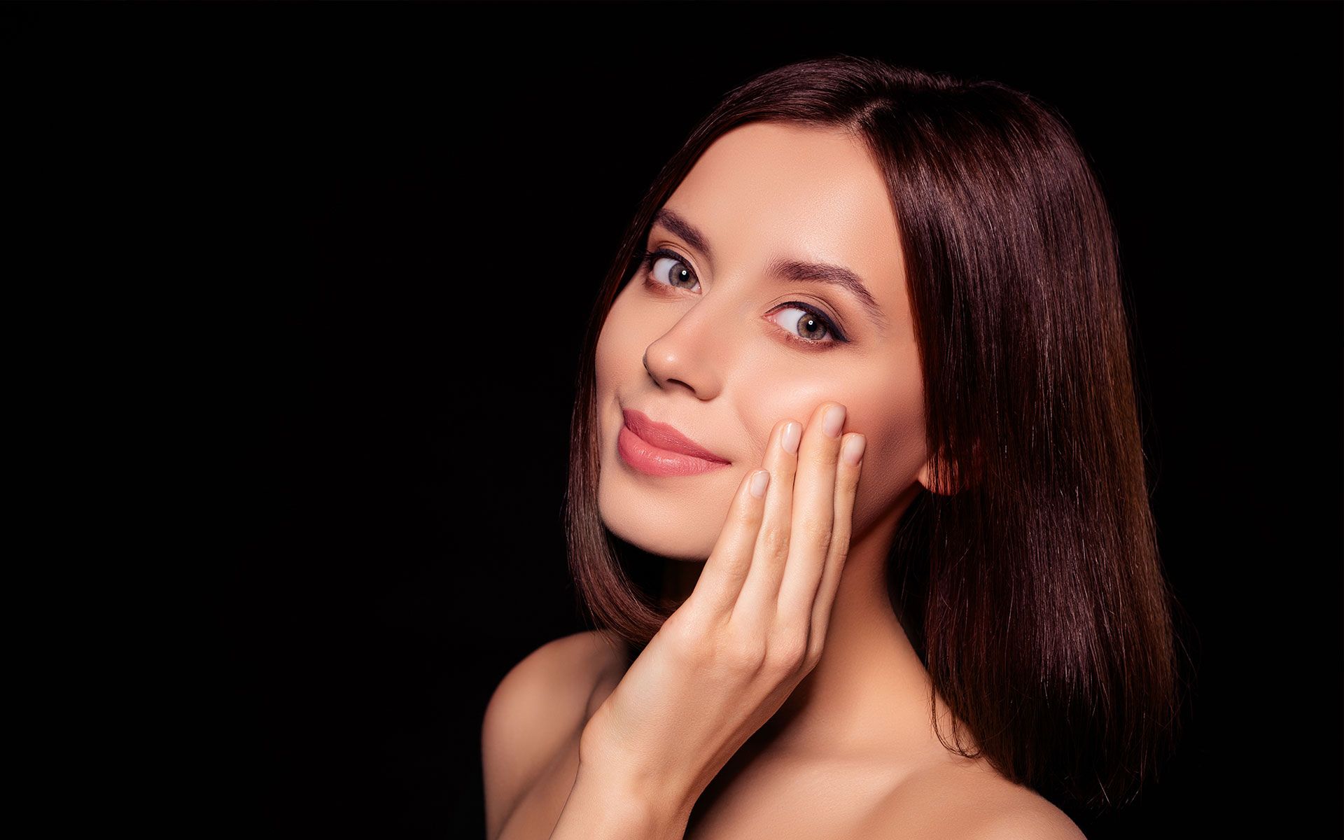 Woman with smooth skin, hand on cheek, smiling, brown hair, black background.
