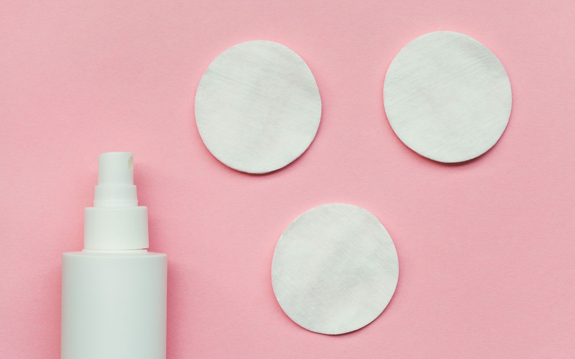 White spray bottle and three cotton pads on a pink background.