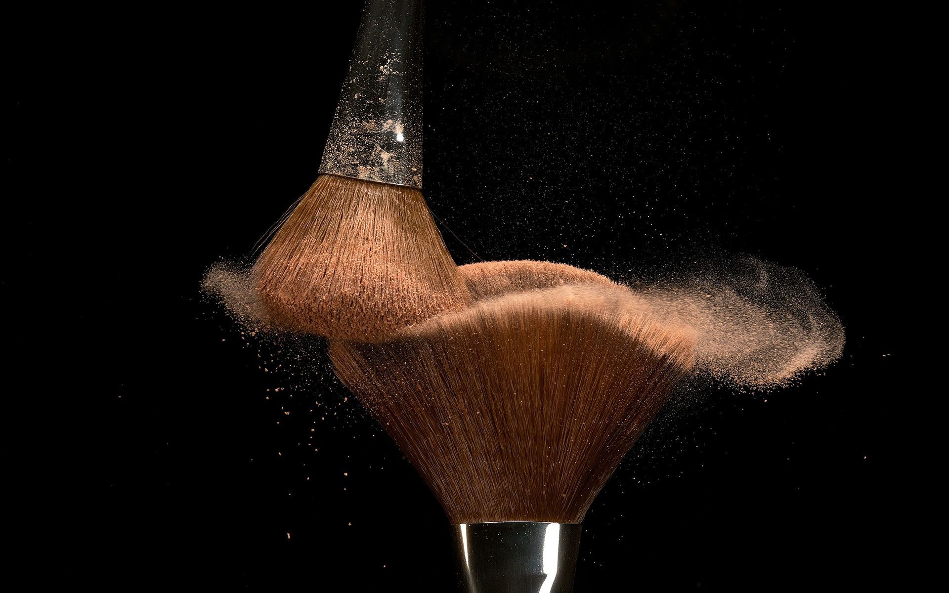 Two makeup brushes colliding, causing a cloud of brown powder to disperse against a black background.