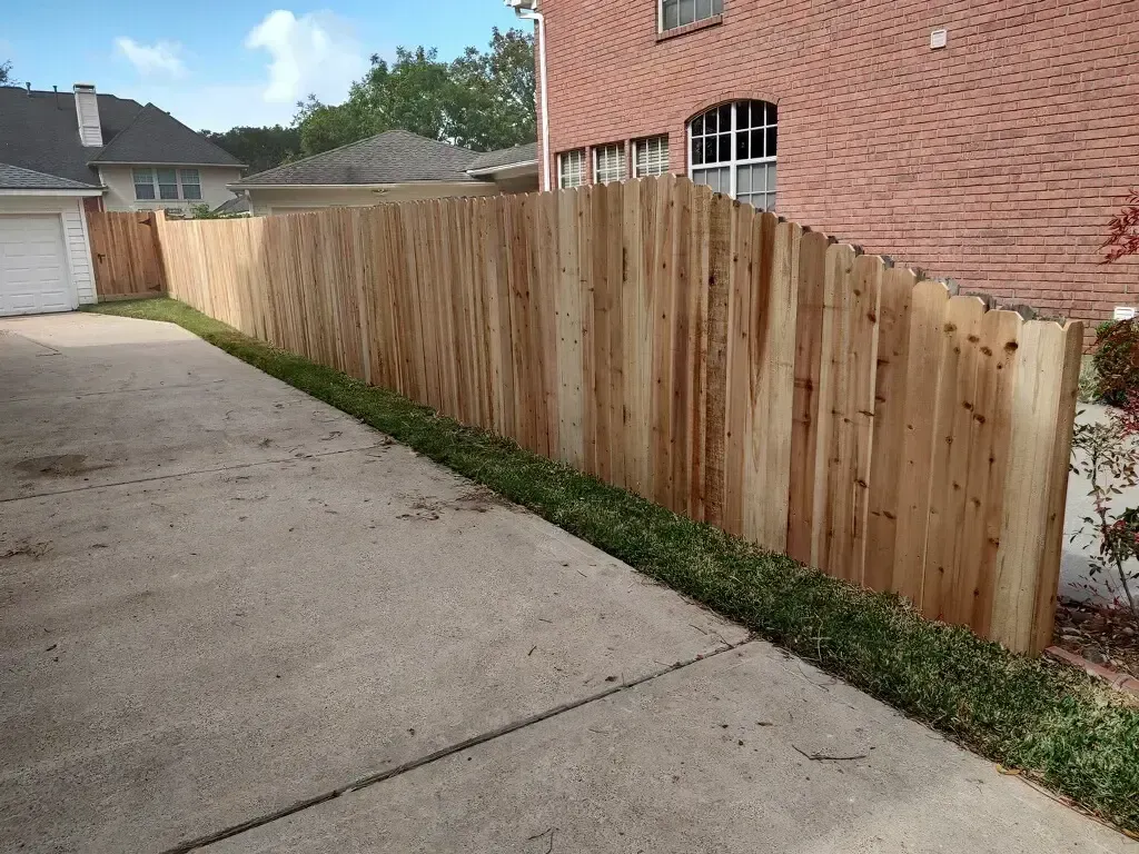 A wooden fence is along the side of a driveway next to a brick house.