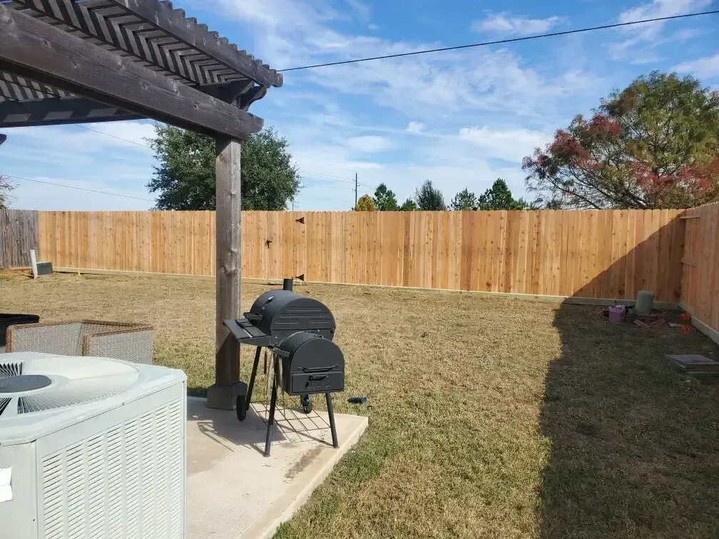 A backyard with a wooden fence , a grill , and an air conditioner.