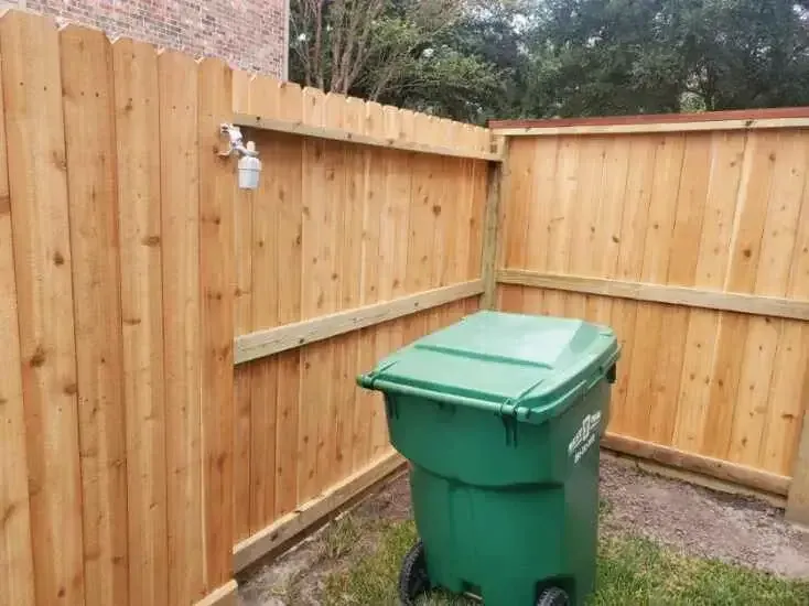 A green trash can is sitting in front of a wooden fence.