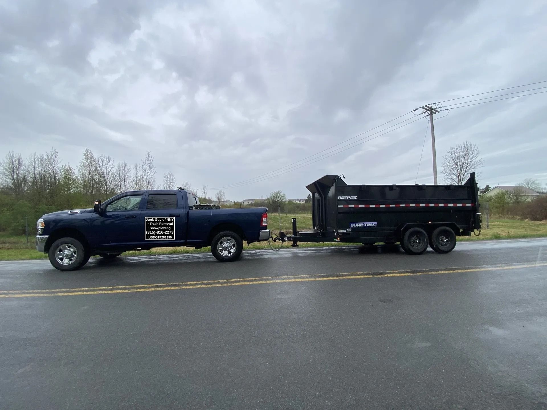 Dark blue pickup truck towing a black dump trailer on a wet road. Overcast sky.