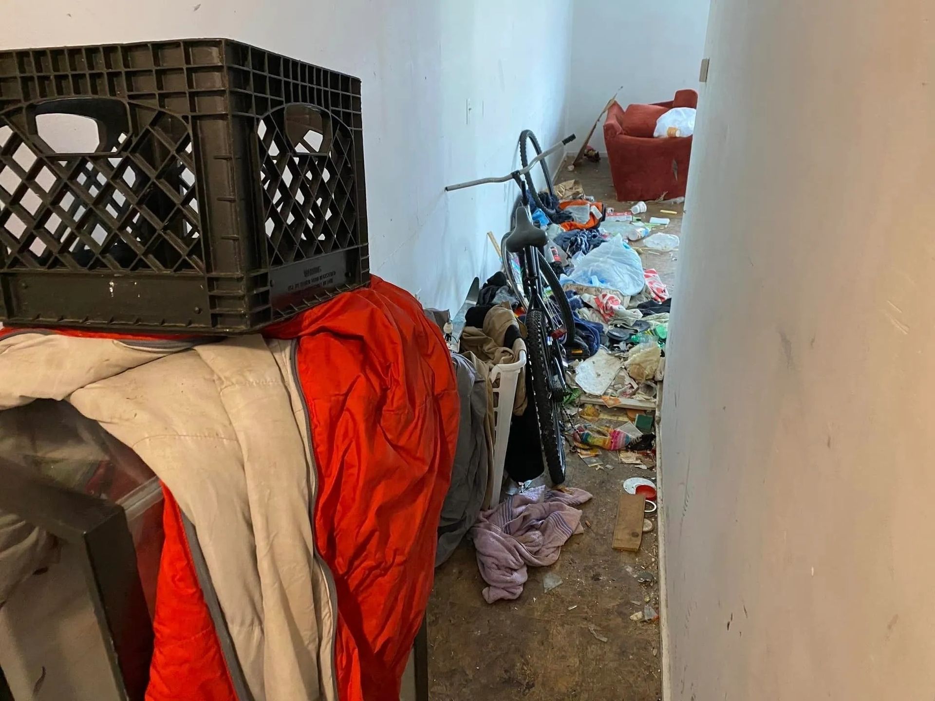 Room with a black crate, red fabric, and a pile of debris, including a bicycle and other items, against a white wall.