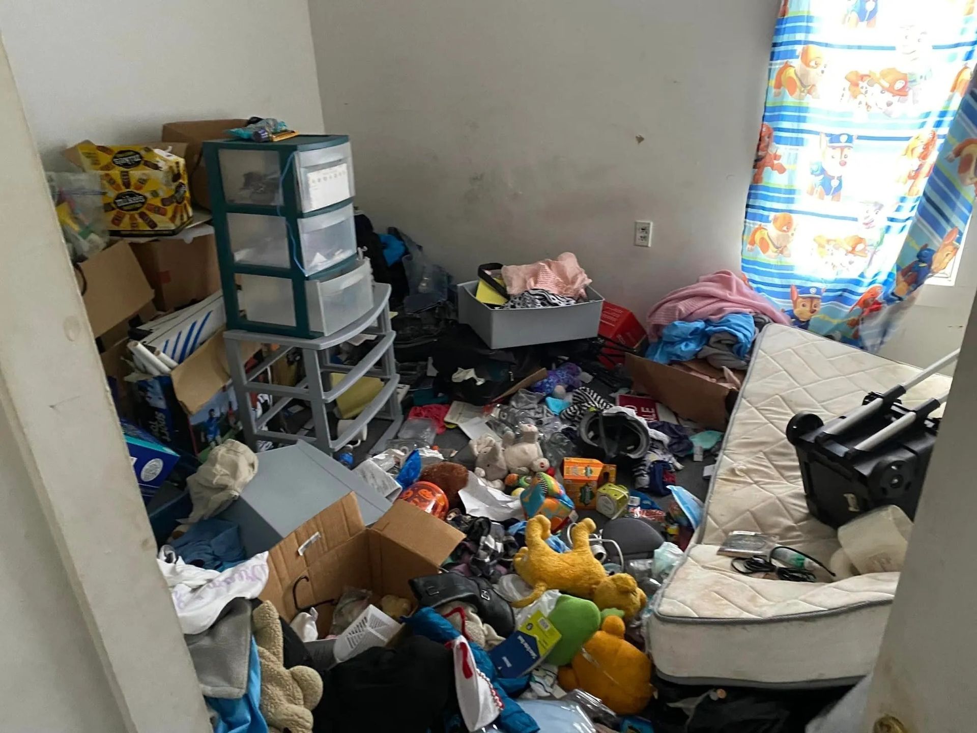 Cluttered room with boxes, clothes, toys, and a mattress on the floor.