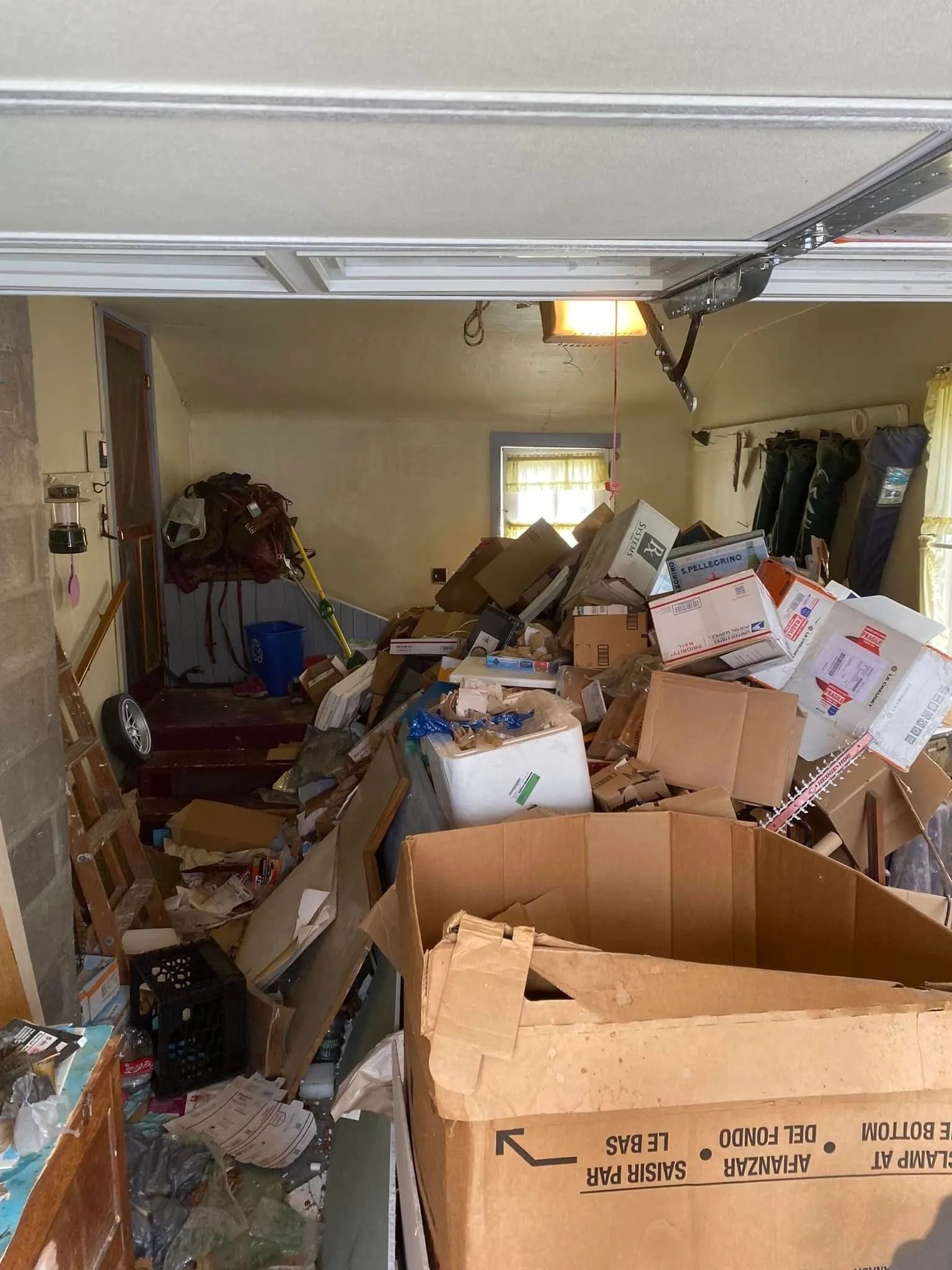 Cluttered garage filled with boxes, furniture, and debris. Sunlight streams in from the back window.