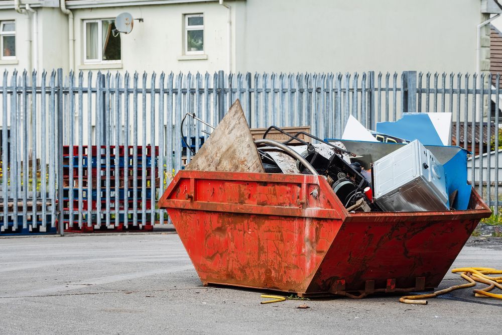 Red dumpster overflowing with scrap metal, wood, and other debris, near a metal fence and building.