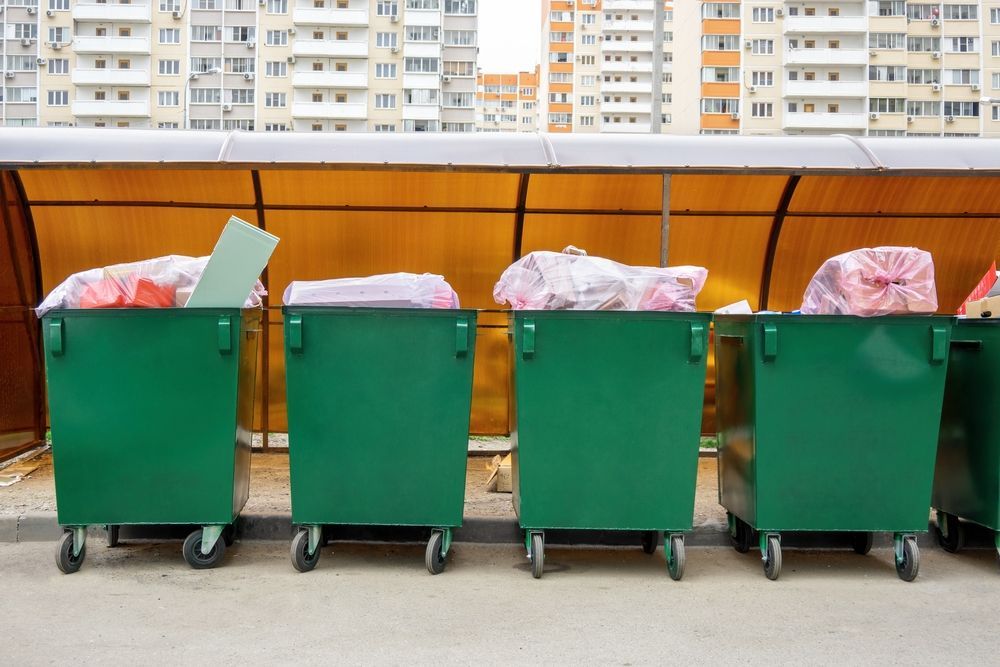 Green dumpsters overflowing with trash beneath a yellow awning, with an apartment building in the background.