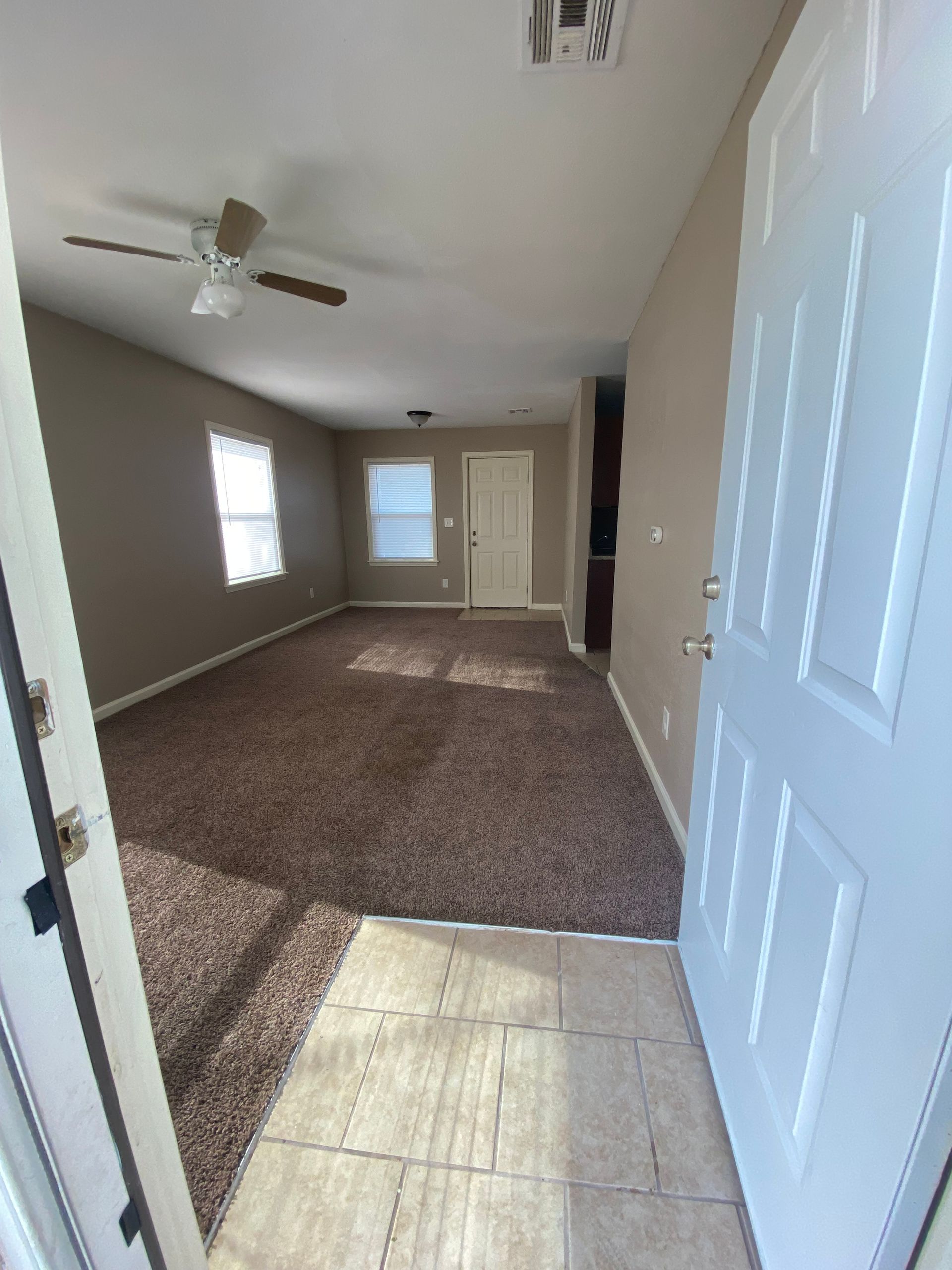 Open doorway into a living room with brown carpet, tan walls, and a ceiling fan.