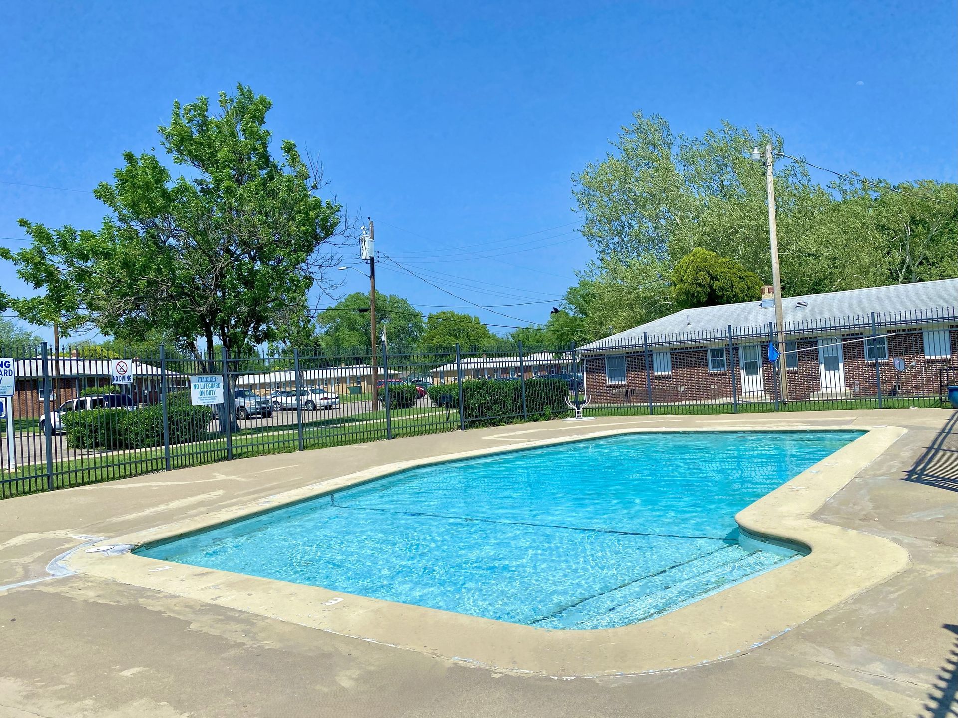Swimming pool surrounded by a fence, with buildings and trees in the background under a blue sky.