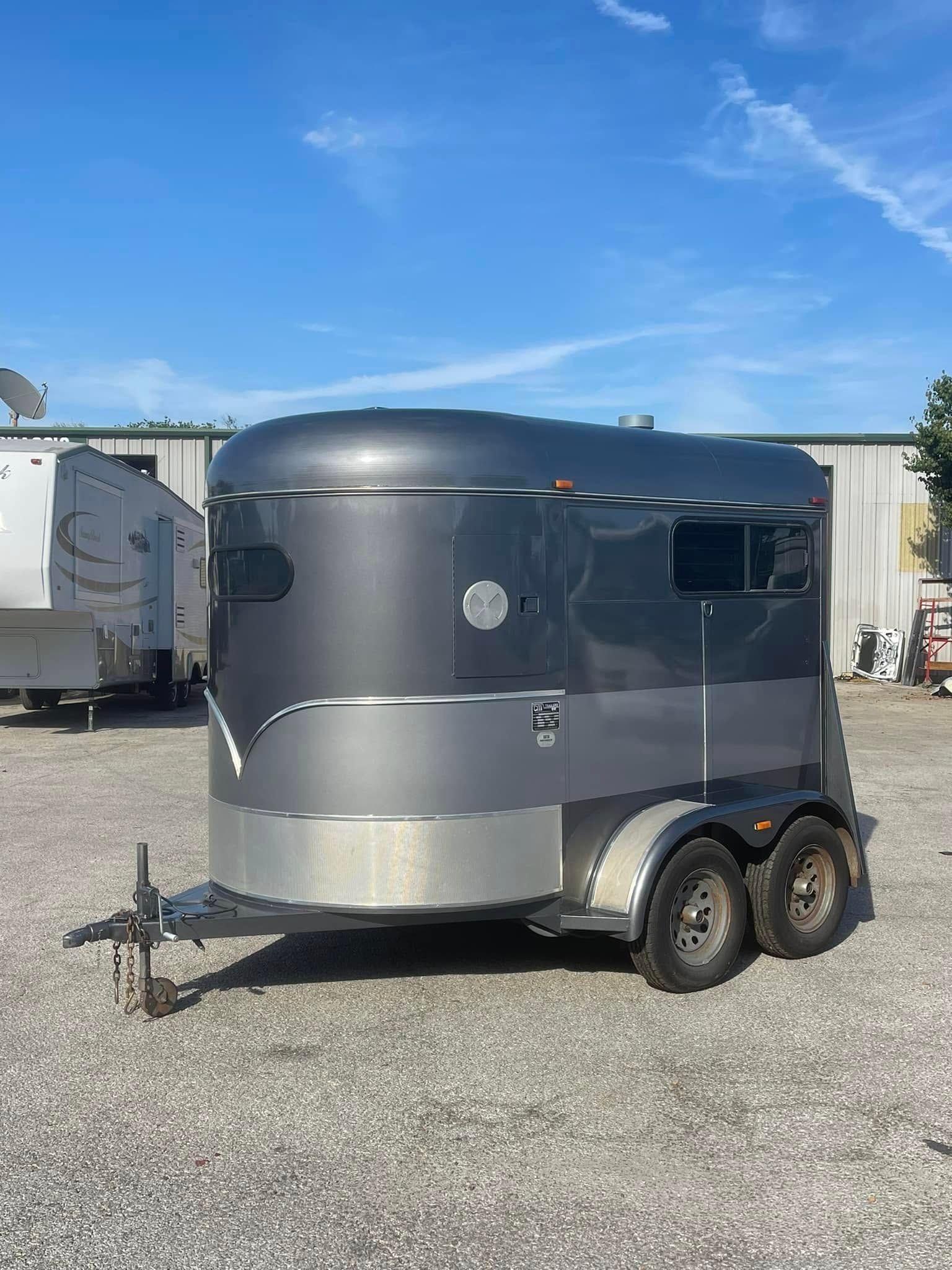 Beige RV trailer with black trim and steps parked outdoors among trees.