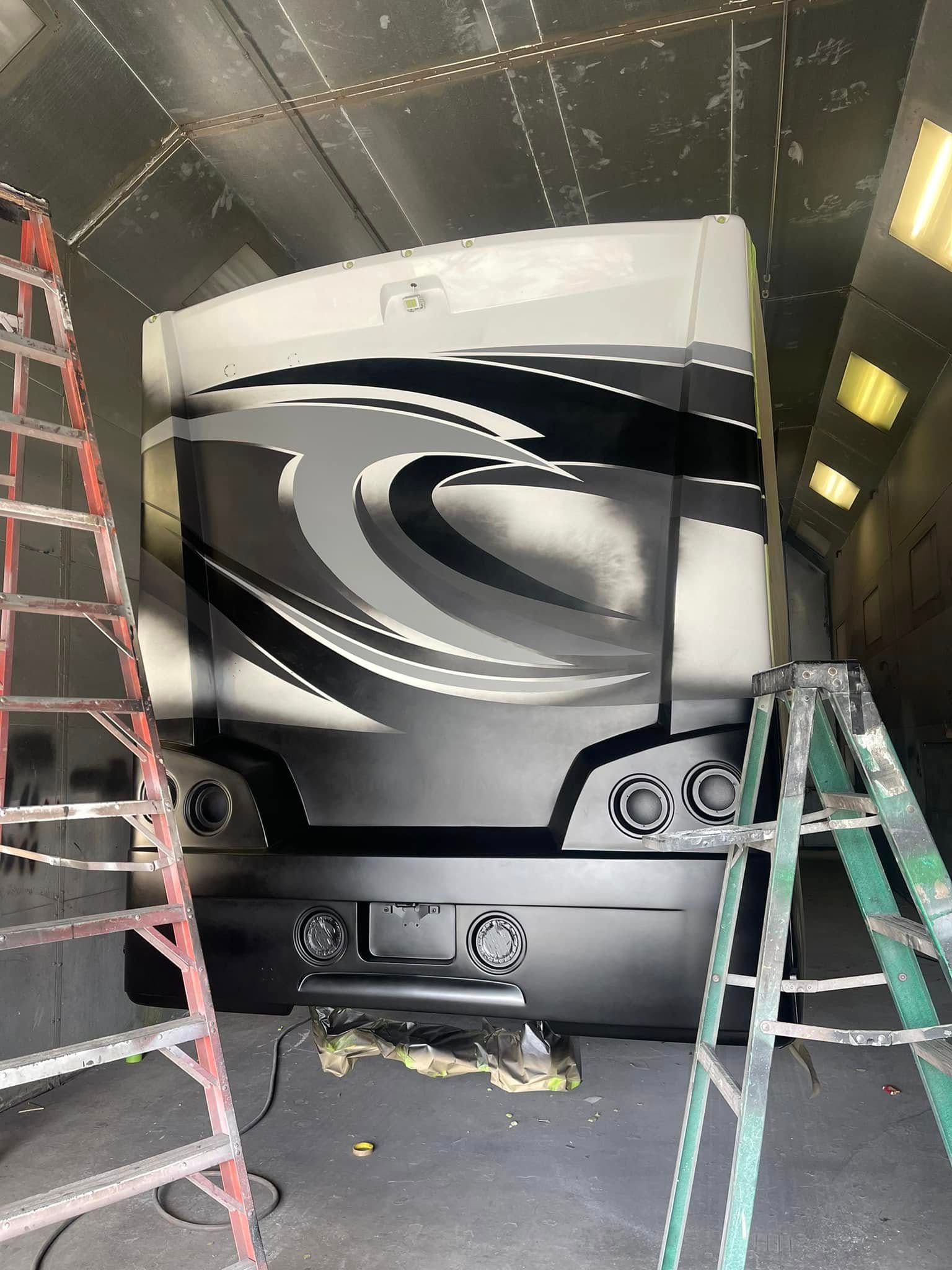 Black and gray RV bus with front panel removed in a workshop, exposing mechanical components and a jack underneath
