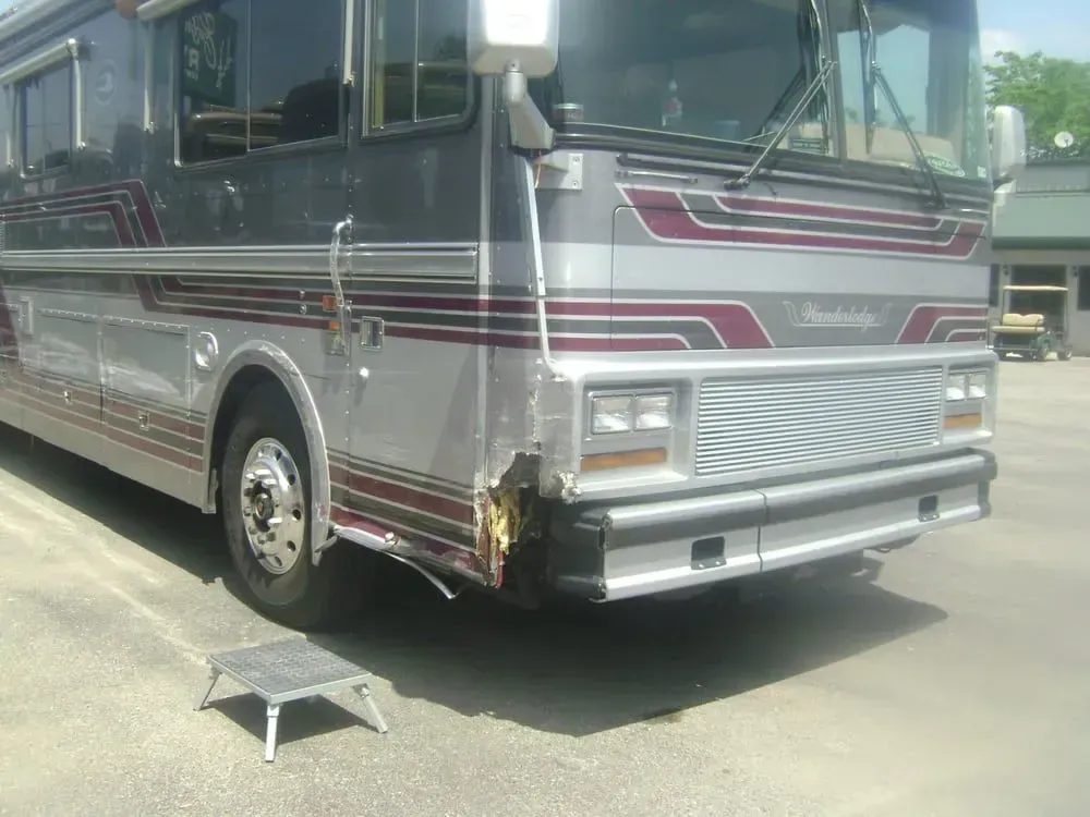 Silver and maroon bus with front damage parked on pavement next to a wheel chock