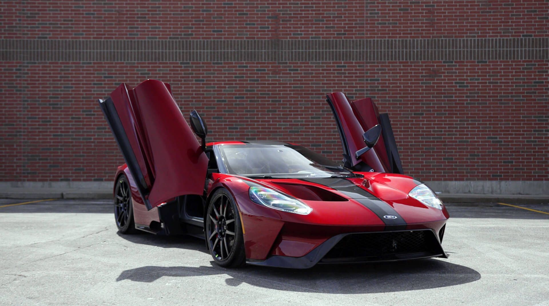 Red sports car with scissor doors open, parked on pavement in front of a brick wall.