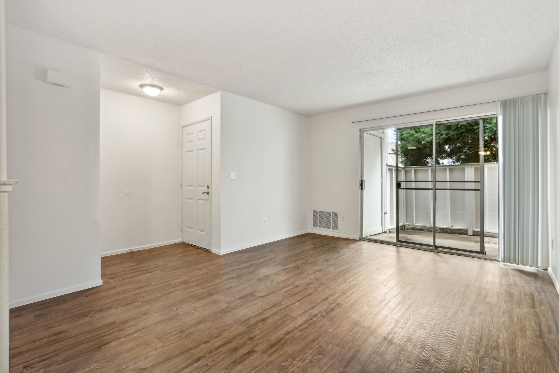 Empty living room with wood flooring, white walls, and a sliding glass door to a small balcony.