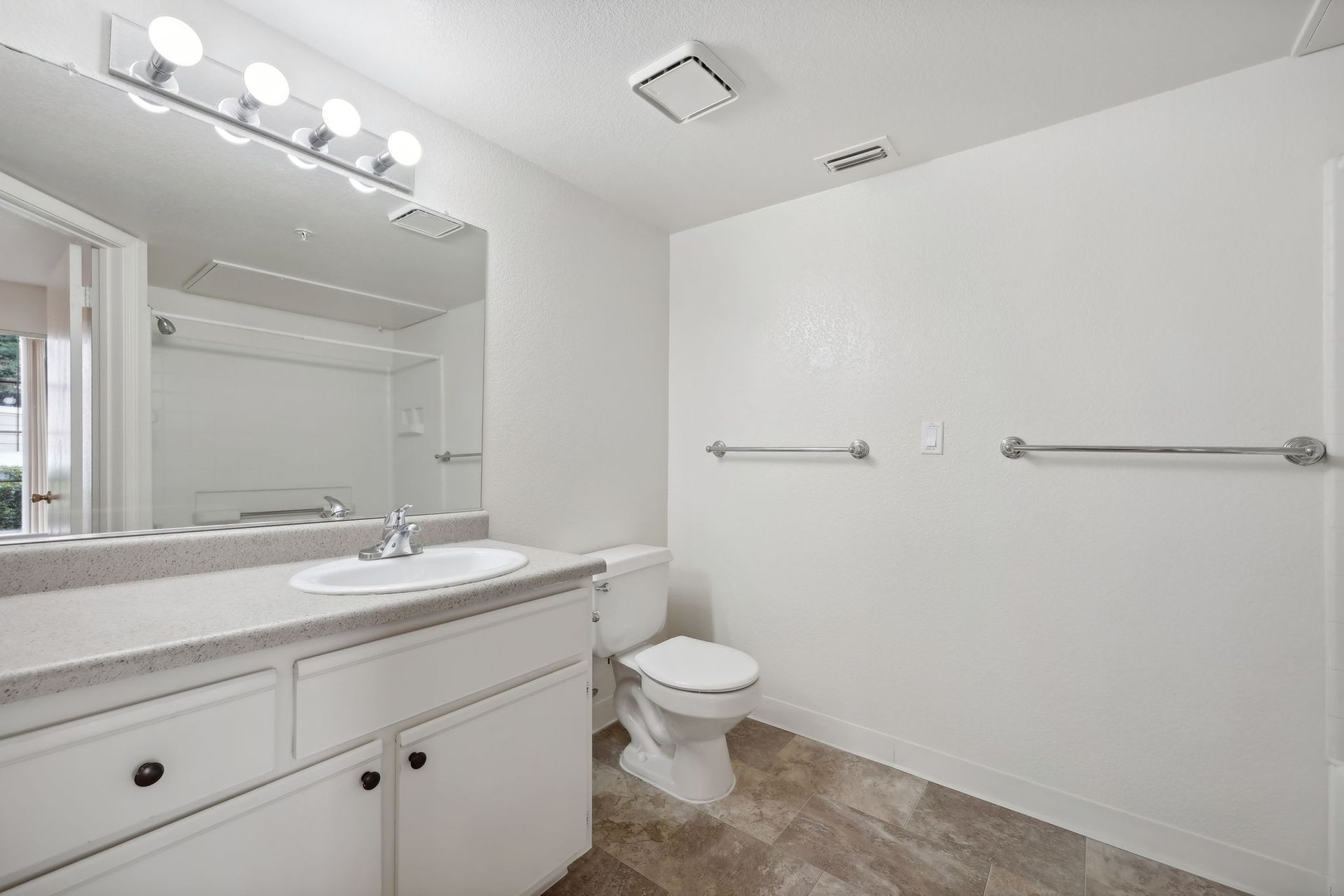 Bathroom with white walls, vanity, toilet, and towel racks. Grayish floor and ceiling vent.