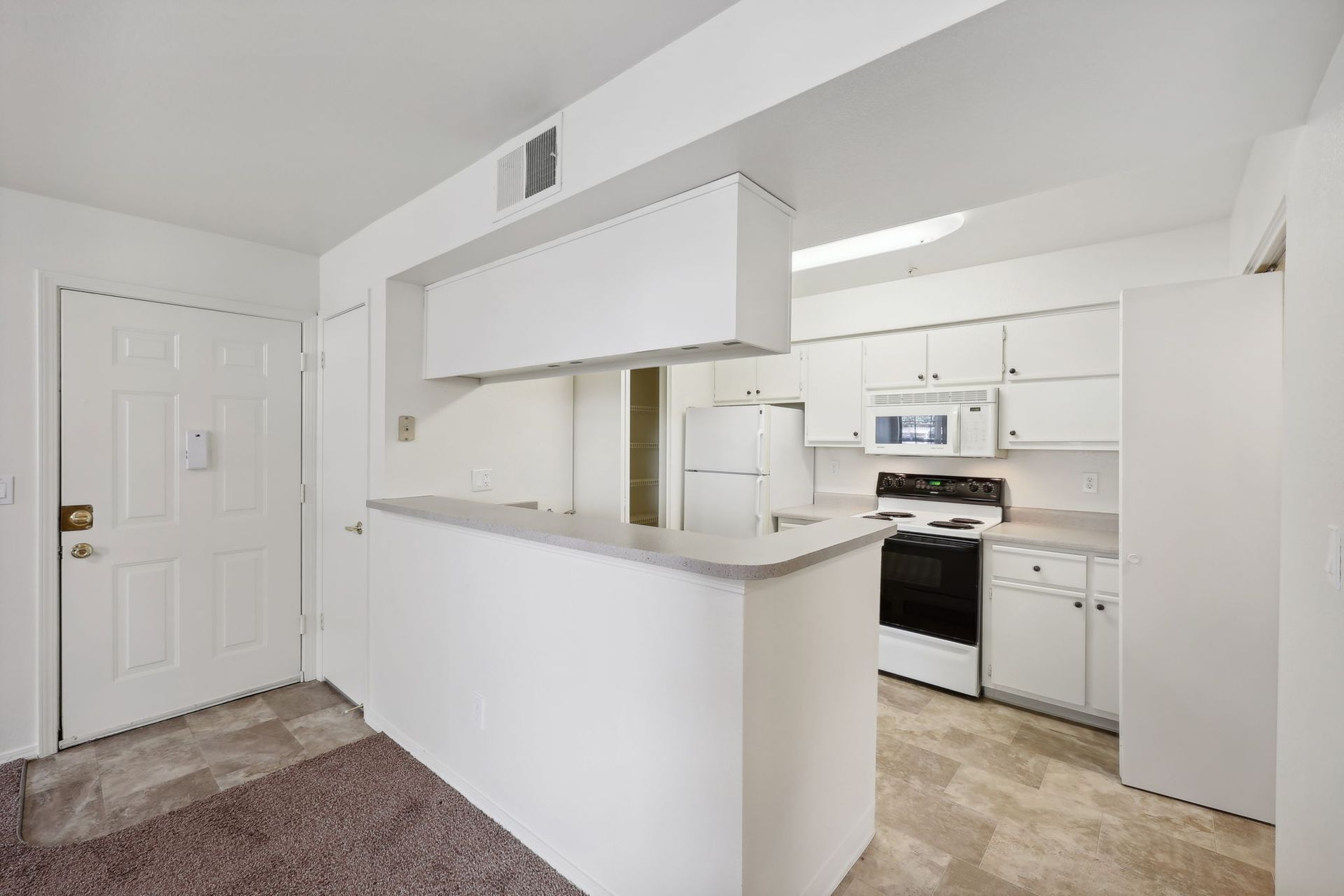 Interior view of a small apartment entryway leading to a white kitchen with appliances and a breakfast bar.
