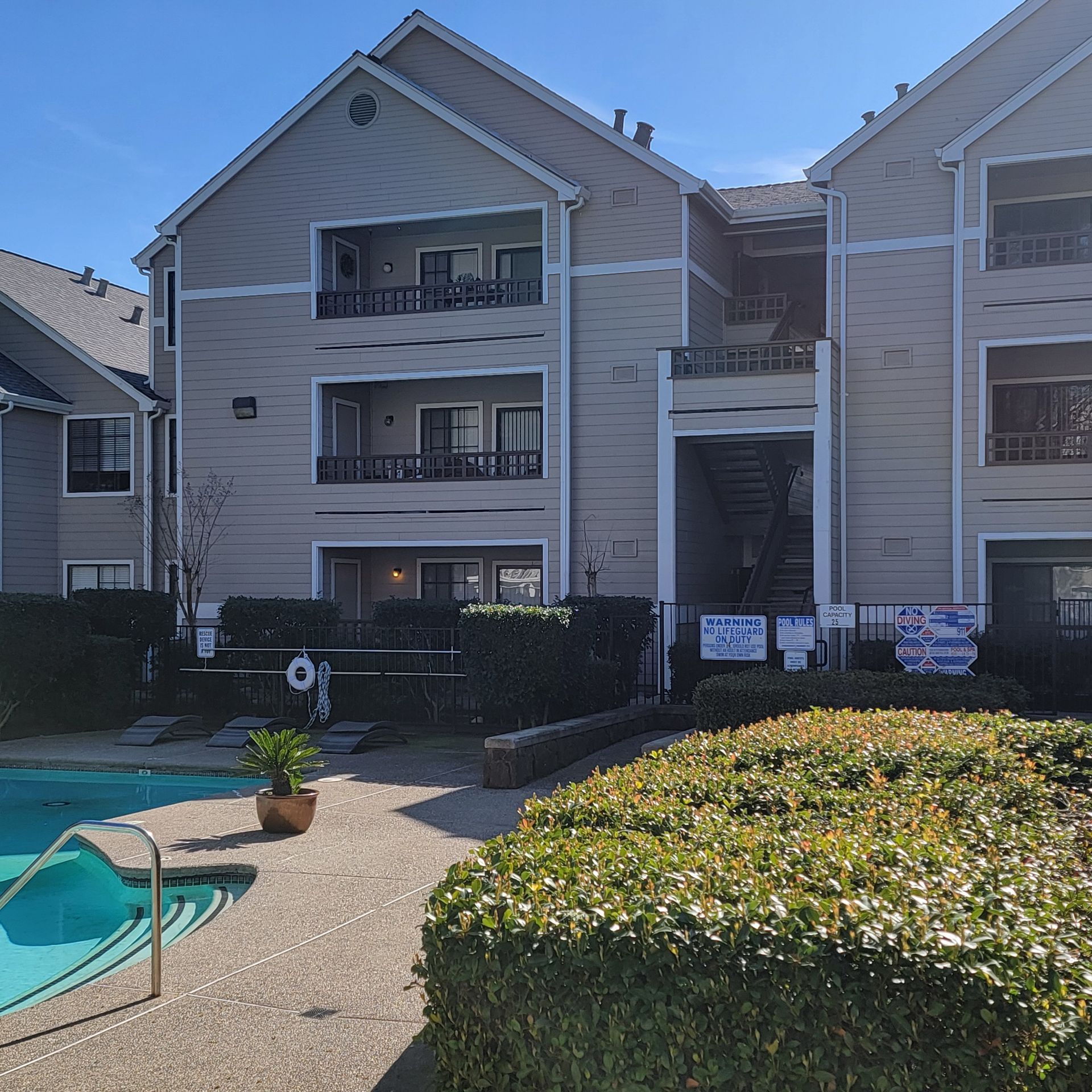 Apartment building with pool. Gray exterior, balconies, and shrubbery in foreground.