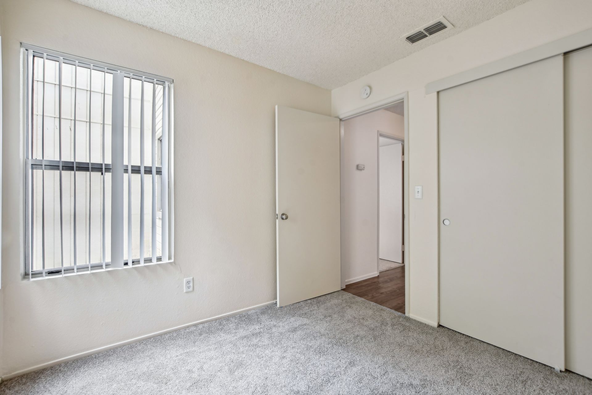 Empty bedroom with a window, white walls, carpet, and sliding closet doors.