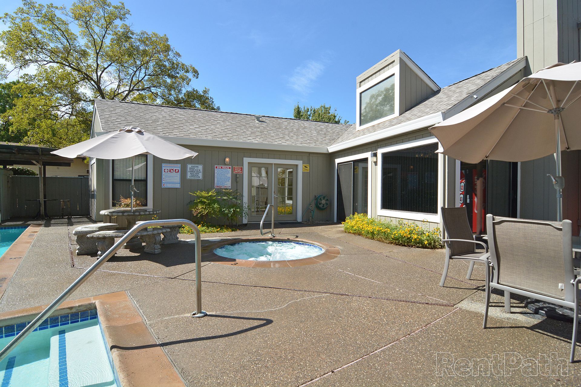 Outdoor area with a pool, spa, seating, and building under a sunny sky.
