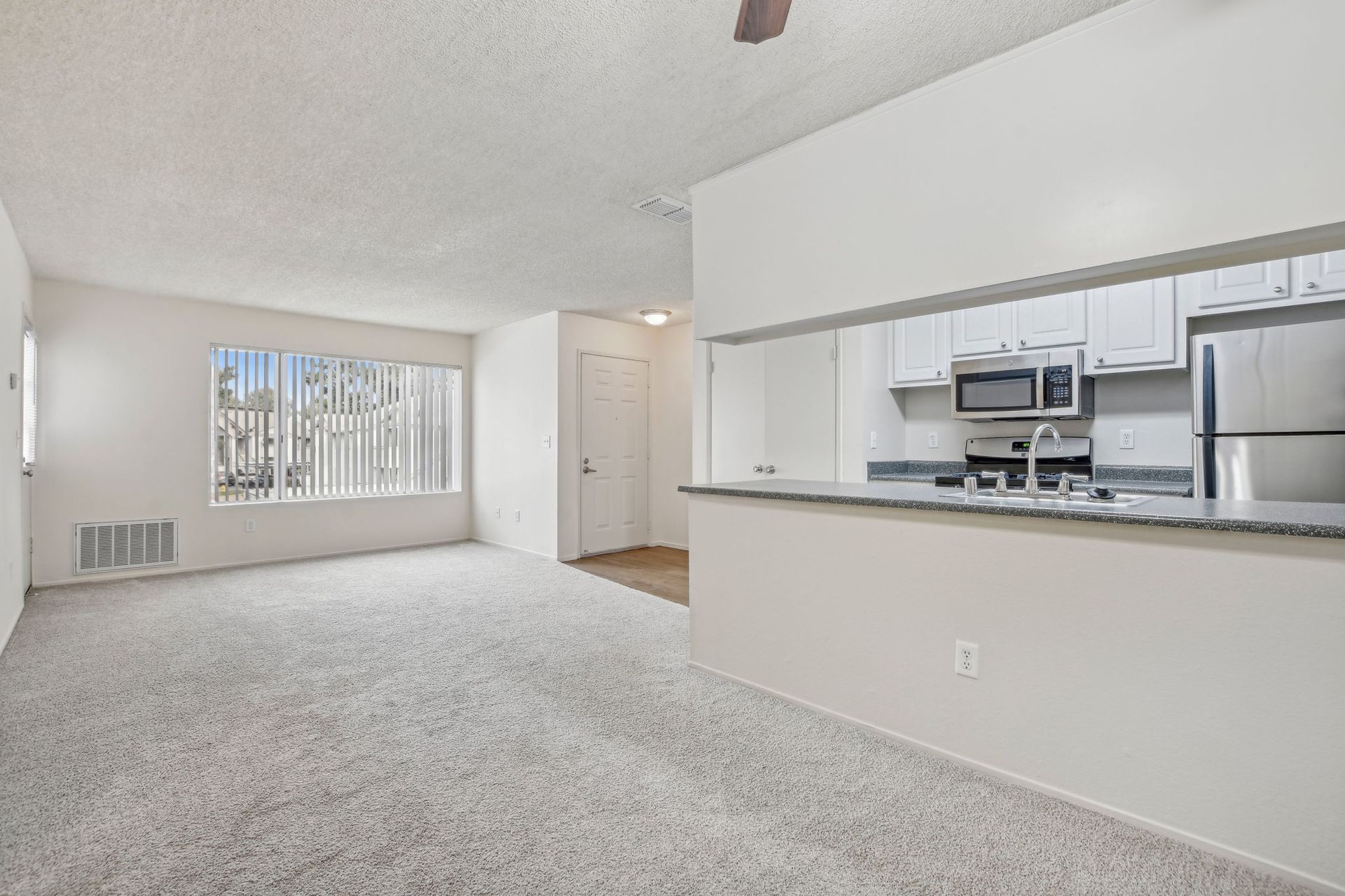 Empty apartment living room with open kitchen, light colors, and carpet flooring.