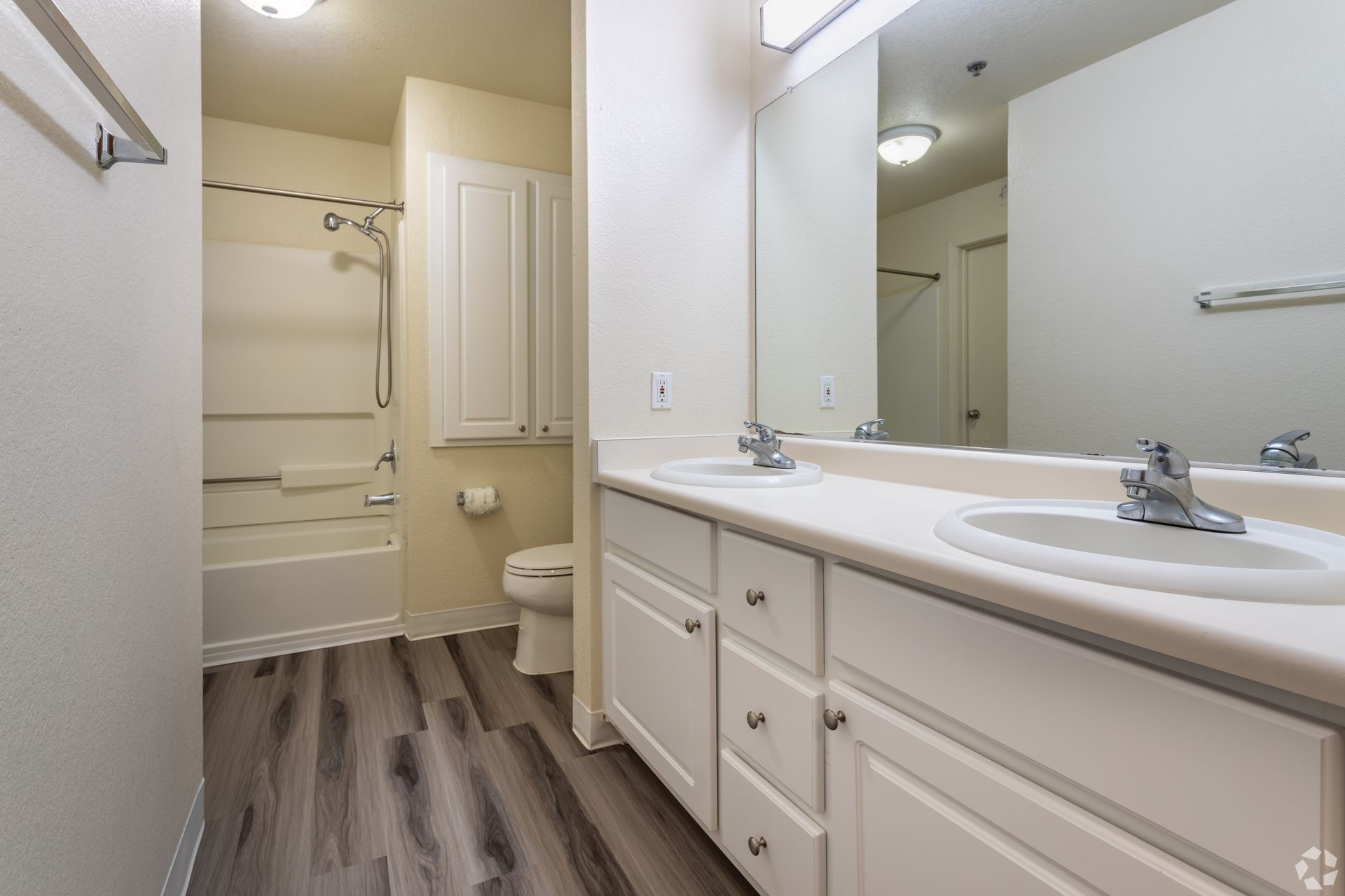 Bathroom with a white vanity, two sinks, and a bathtub with a showerhead. Wood-look flooring.