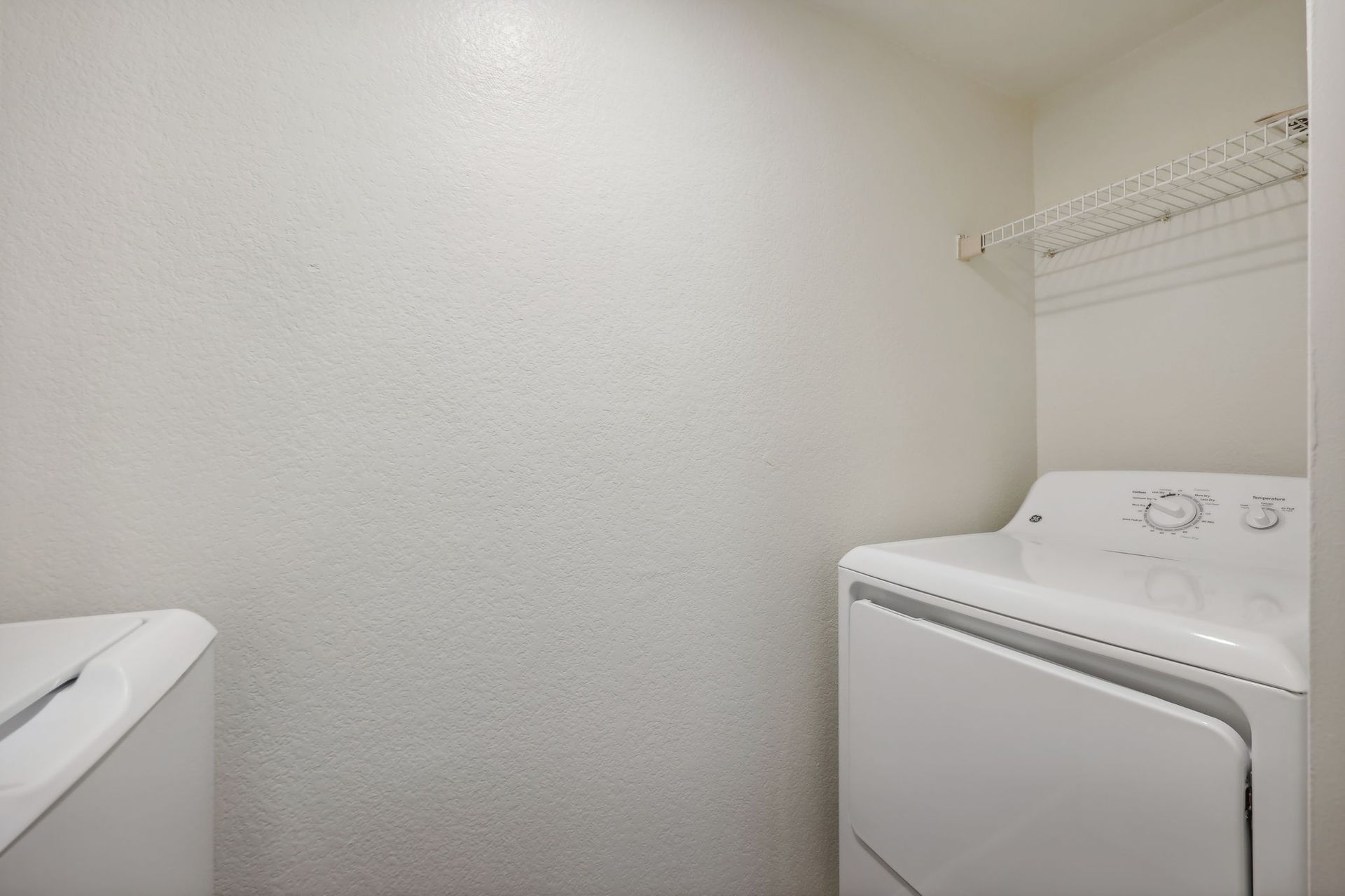Laundry room with white washer and dryer, wire shelf, and textured white walls.
